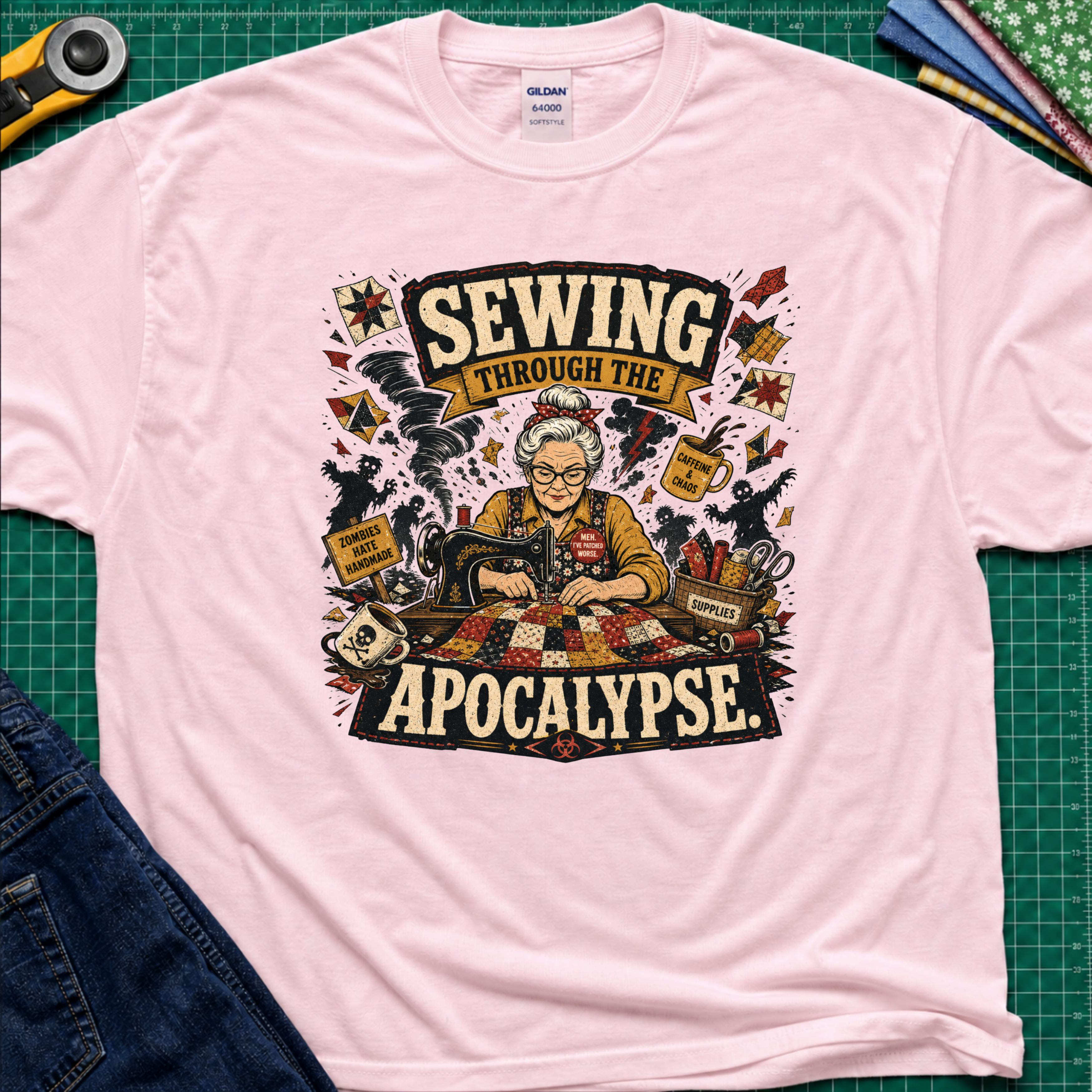 A light pink Gildan t-shirt laid flat on a green cutting mat, featuring a detailed graphic of a woman sewing with the text 'SEWING THROUGH THE APOCALYPSE' and various retro sewing and apocalypse-themed illustrations.