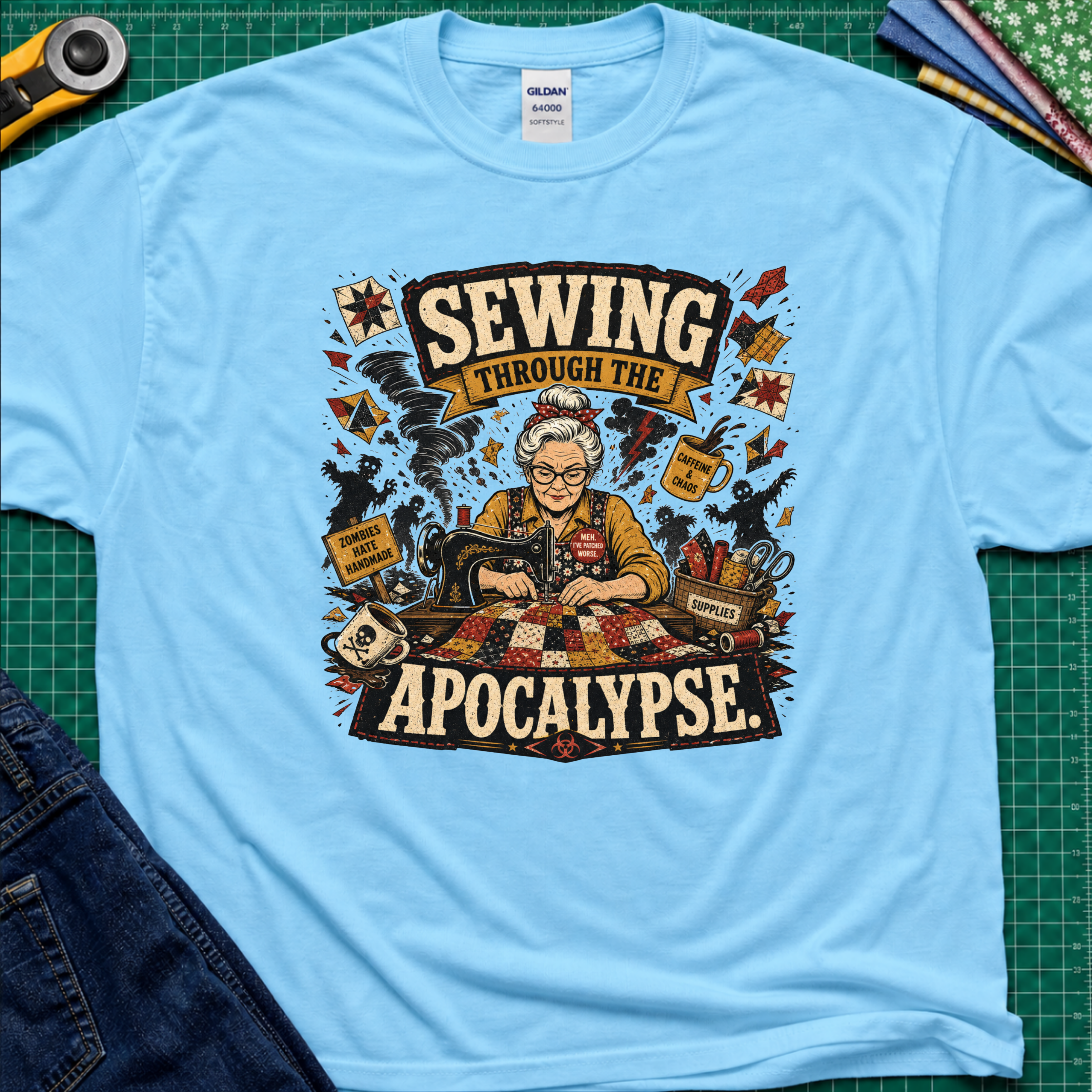 A sky blue Gildan t-shirt laid flat on a green cutting mat, featuring a detailed graphic of a woman sewing with the text 'SEWING THROUGH THE APOCALYPSE' and various retro sewing and apocalypse-themed illustrations.