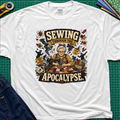 A white Gildan t-shirt laid flat on a green cutting mat, featuring a detailed graphic of a woman sewing with the text 'SEWING THROUGH THE APOCALYPSE' and various retro sewing and apocalypse-themed illustrations.