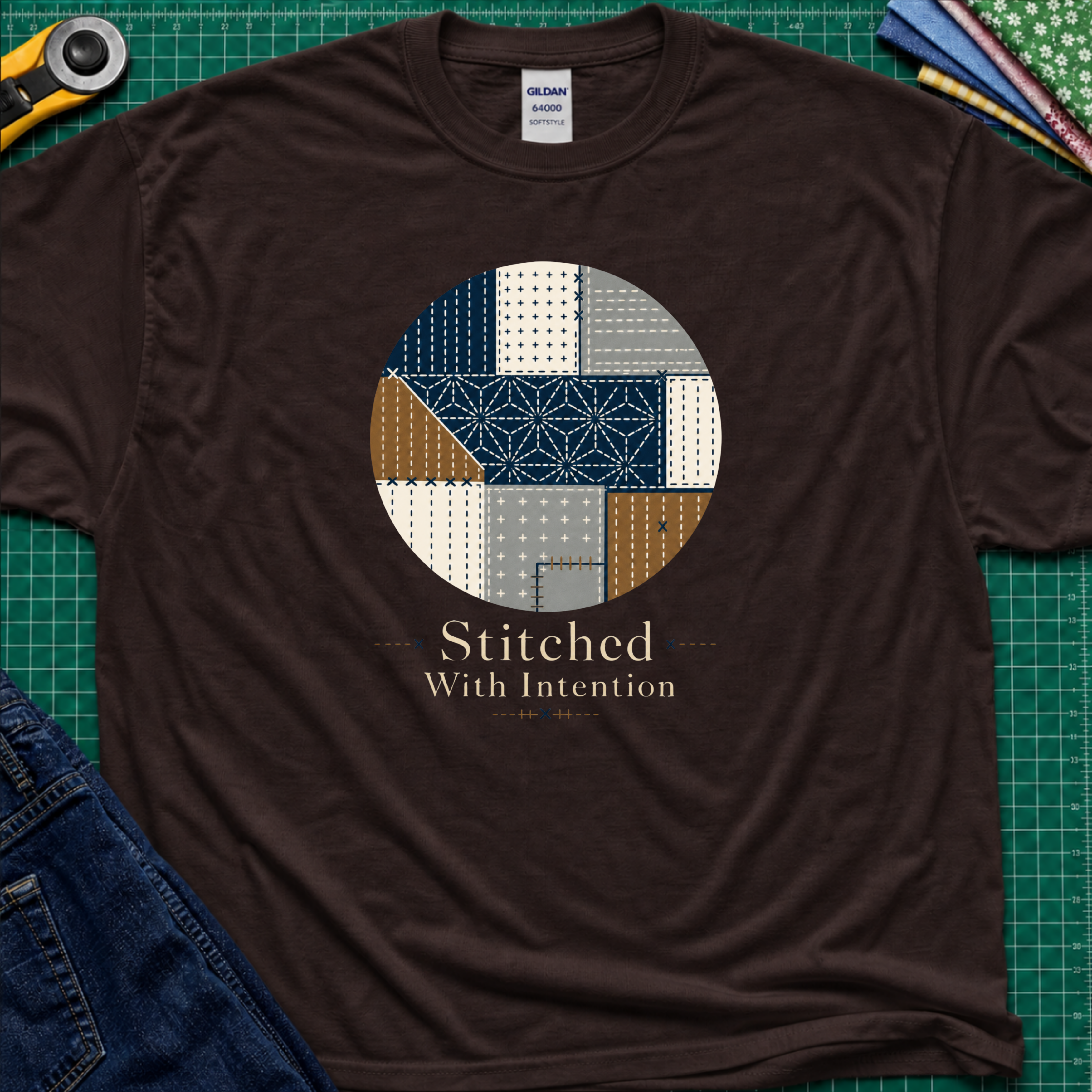 A dark chocolate Gildan t-shirt featuring a circular sashiko graphic in navy, beige, gray, and brown with the text 'Stitched With Intention' printed below. The shirt lays flat on a green cutting mat with a rotary cutter, fabric stack and blue jeans partially visible in the corners.