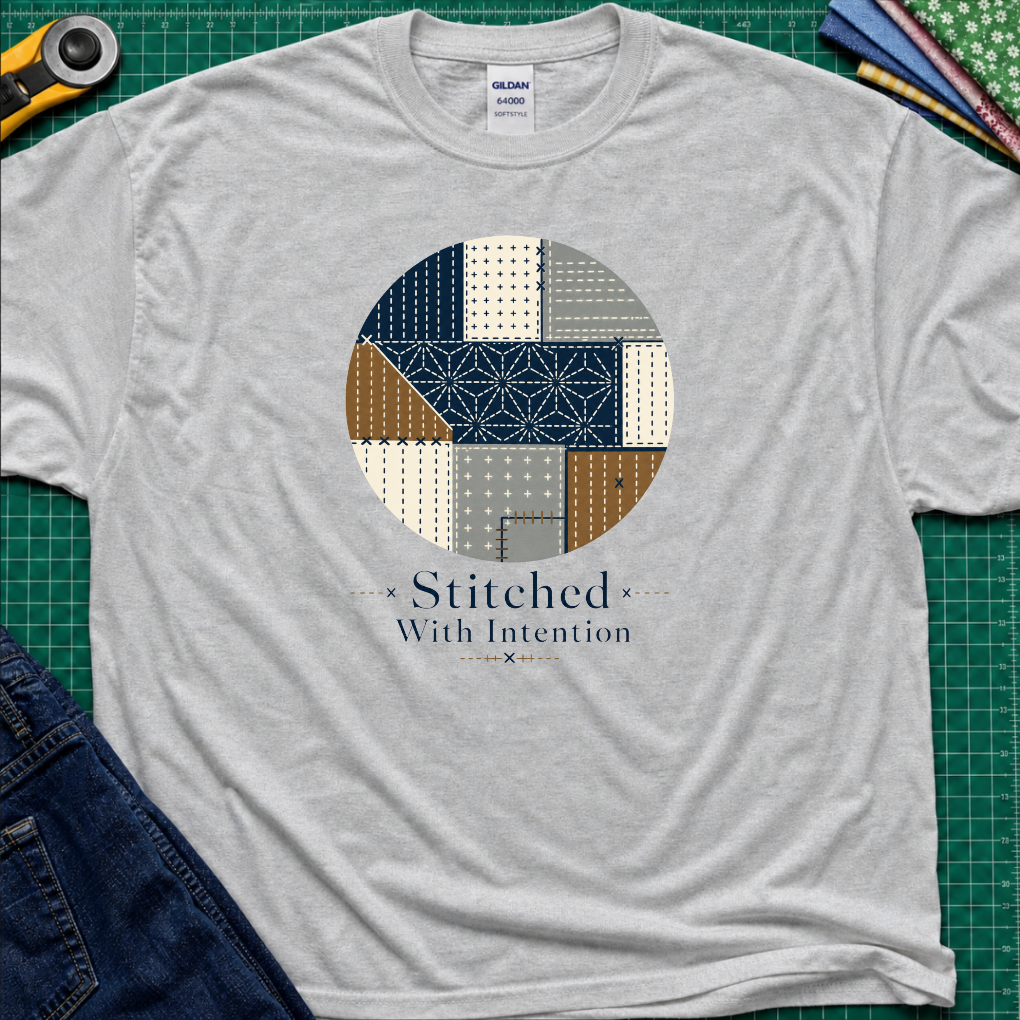 A sports grey Gildan t-shirt featuring a circular sashiko graphic in navy, beige, gray, and brown with the text 'Stitched With Intention' printed below. The shirt lays flat on a green cutting mat with a rotary cutter, fabric stack and blue jeans partially visible in the corners.
