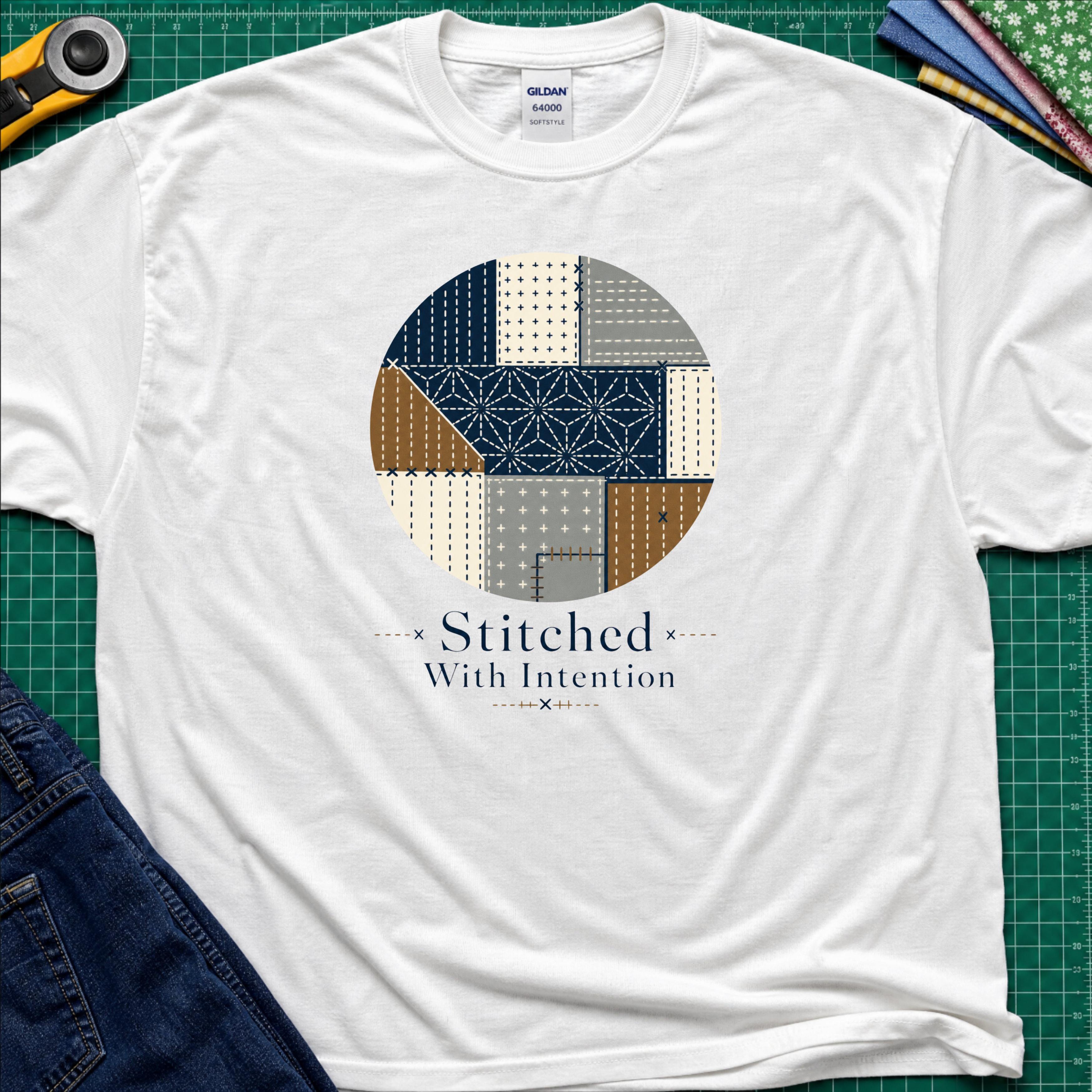 A white Gildan t-shirt featuring a circular sashiko graphic in navy, beige, gray, and brown with the text 'Stitched With Intention' printed below. The shirt lays flat on a green cutting mat with a rotary cutter, fabric stack and blue jeans partially visible in the corners.