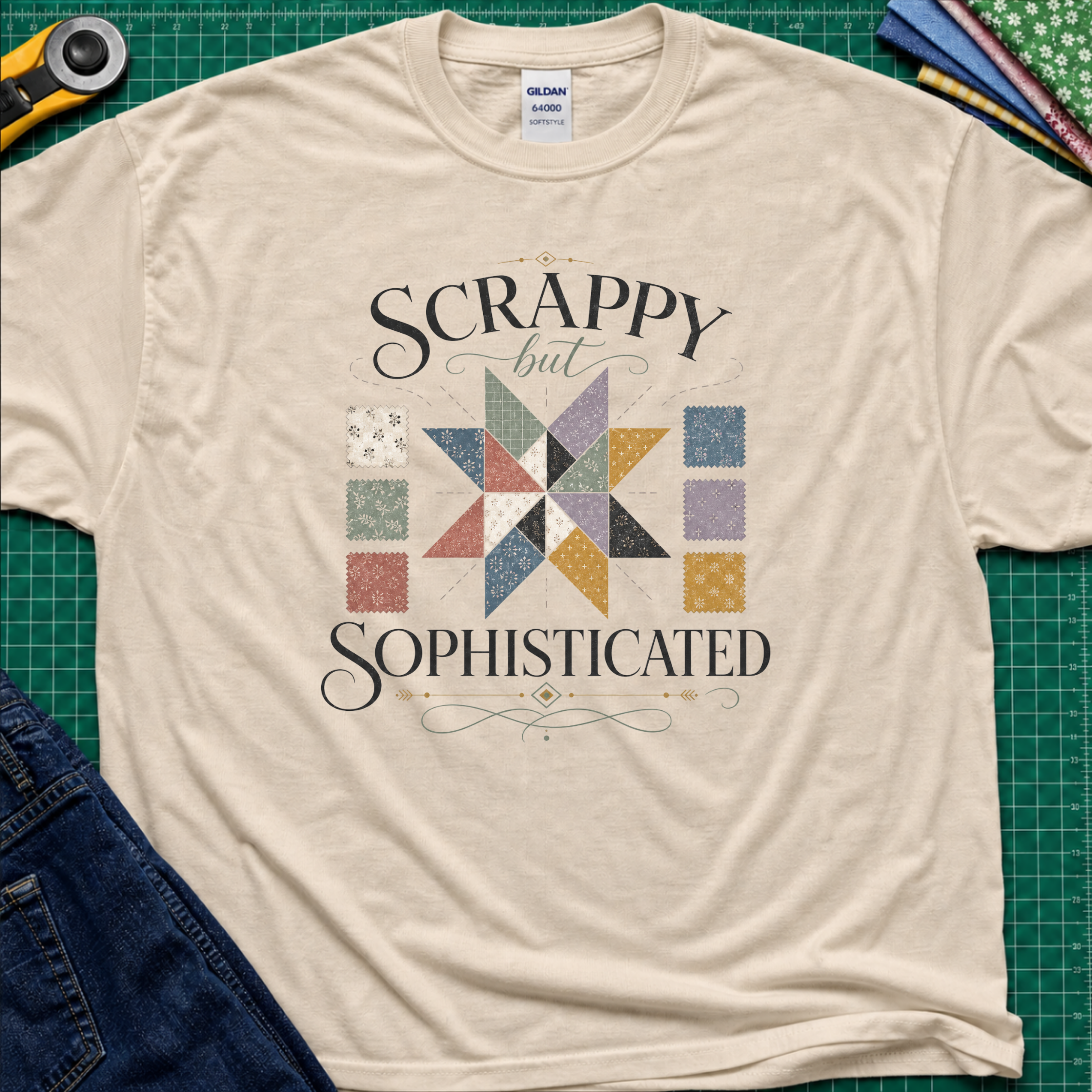A dark cream Gildan t-shirt laid flat on a green cutting mat background, featuring a colorful quilted star graphic and the words 'SCRAPPY but SOPHISTICATED' in black script. A rotary cutter, fabric, and jeans are partially visible as styling props.