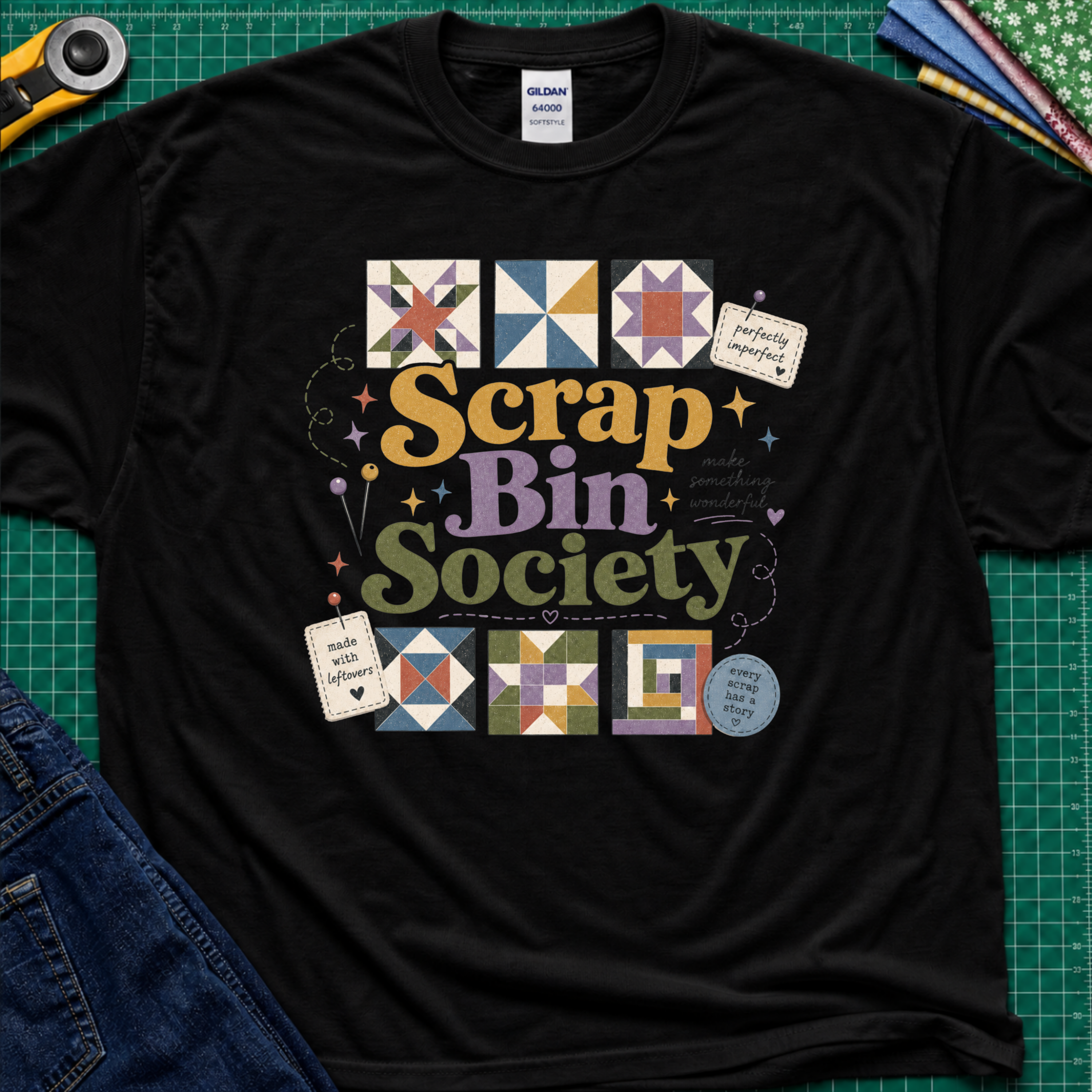 A black Gildan t-shirt featuring a colorful 'Scrap Bin Society' graphic with quilt patterns, laid flat on a green cutting mat with sewing tools and jeans partially visible.
