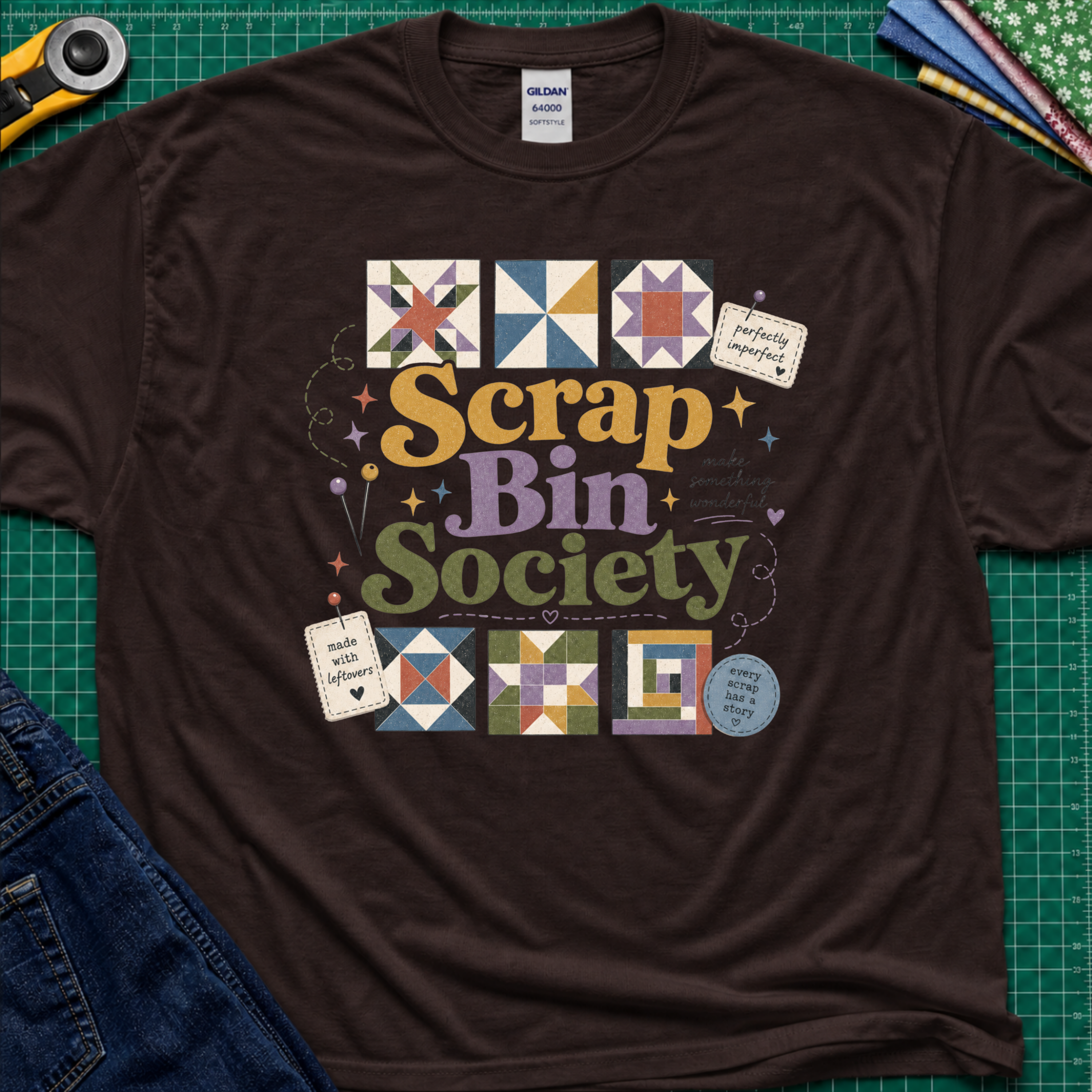 A dark brown Gildan t-shirt with a colorful 'Scrap Bin Society' graphic printed on the front, styled in a flat-lay arrangement on a green cutting mat with sewing tools and denim jeans.