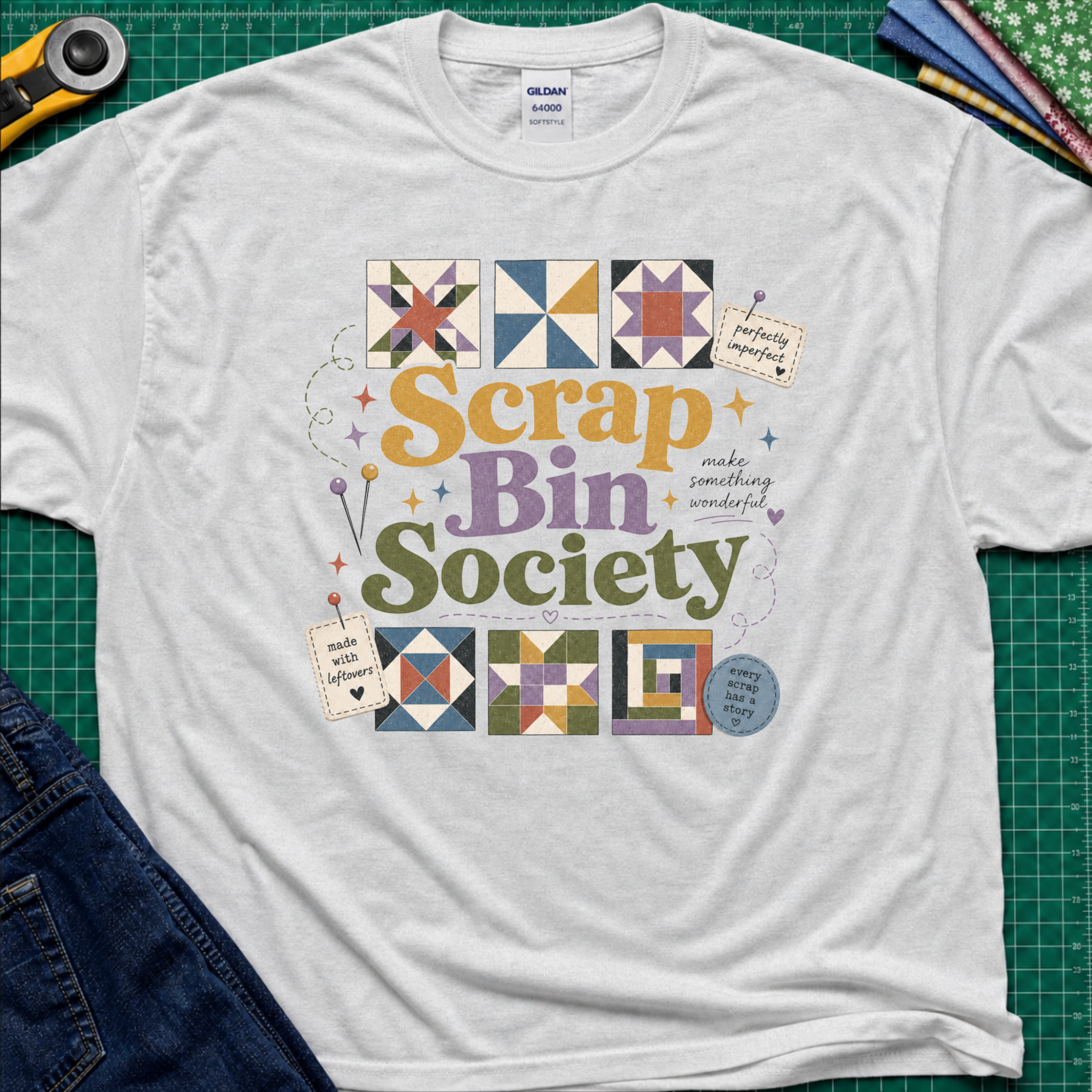 A white Gildan t-shirt featuring a colorful 'Scrap Bin Society' graphic with quilt patterns, laid flat on a green cutting mat with sewing tools and jeans partially visible.