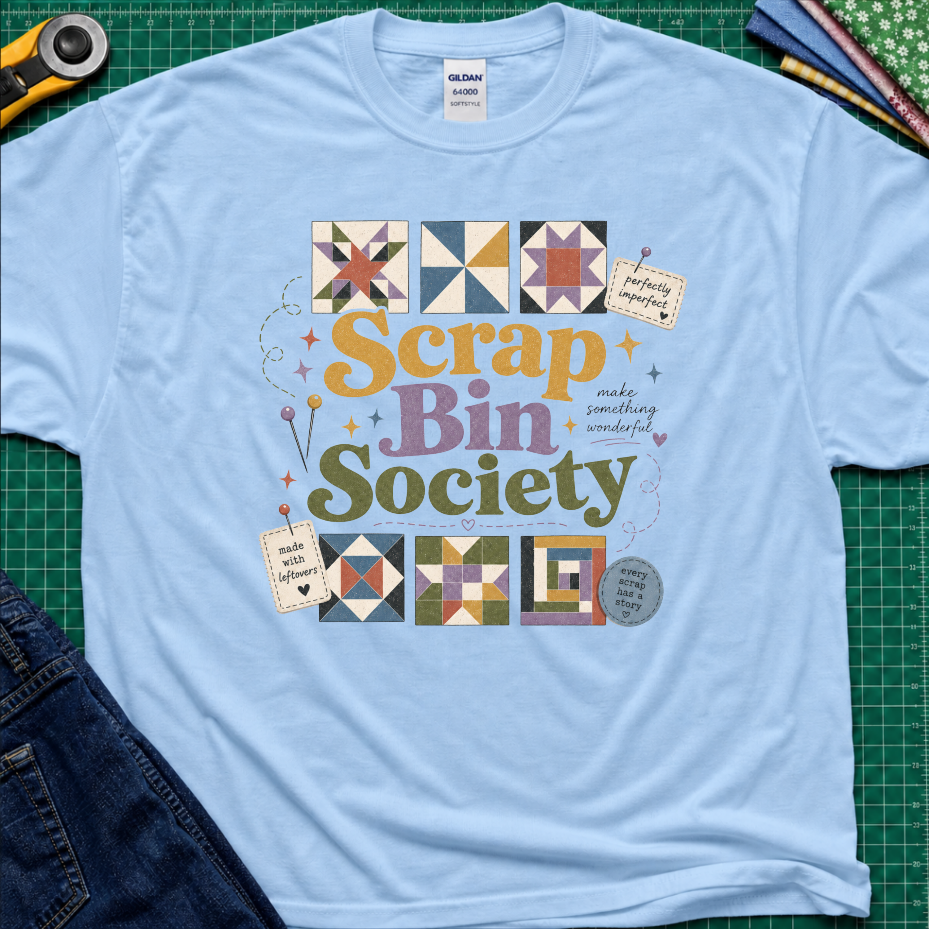 A light blue t-shirt with a colorful 'Scrap Bin Society' graphic printed on the chest, styled in a flat-lay on a green cutting mat with crafting tools like a rotary cutter and jeans visible around it.