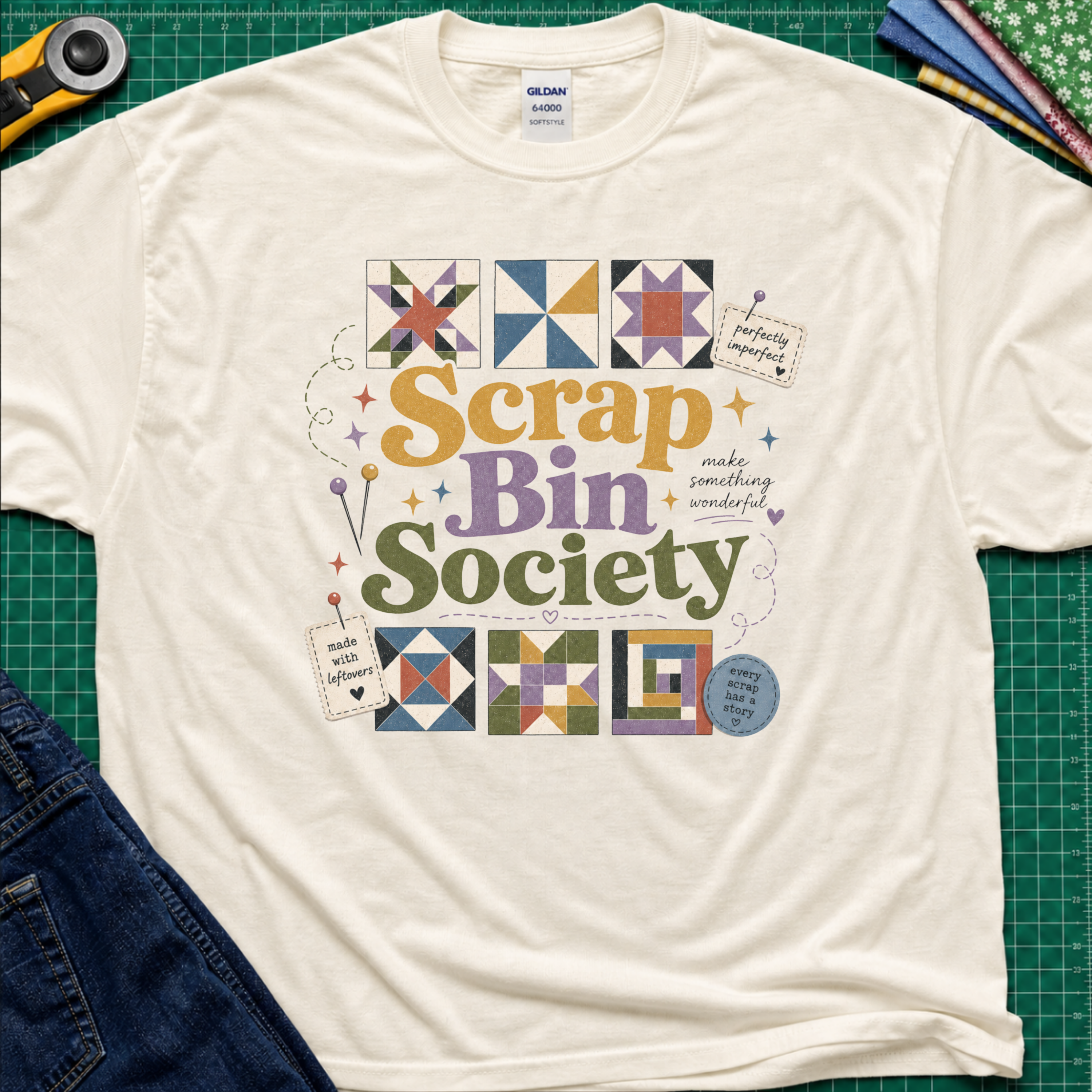 A light cream Gildan t-shirt featuring a colorful 'Scrap Bin Society' graphic with quilt patterns, laid flat on a green cutting mat with sewing tools and jeans partially visible.