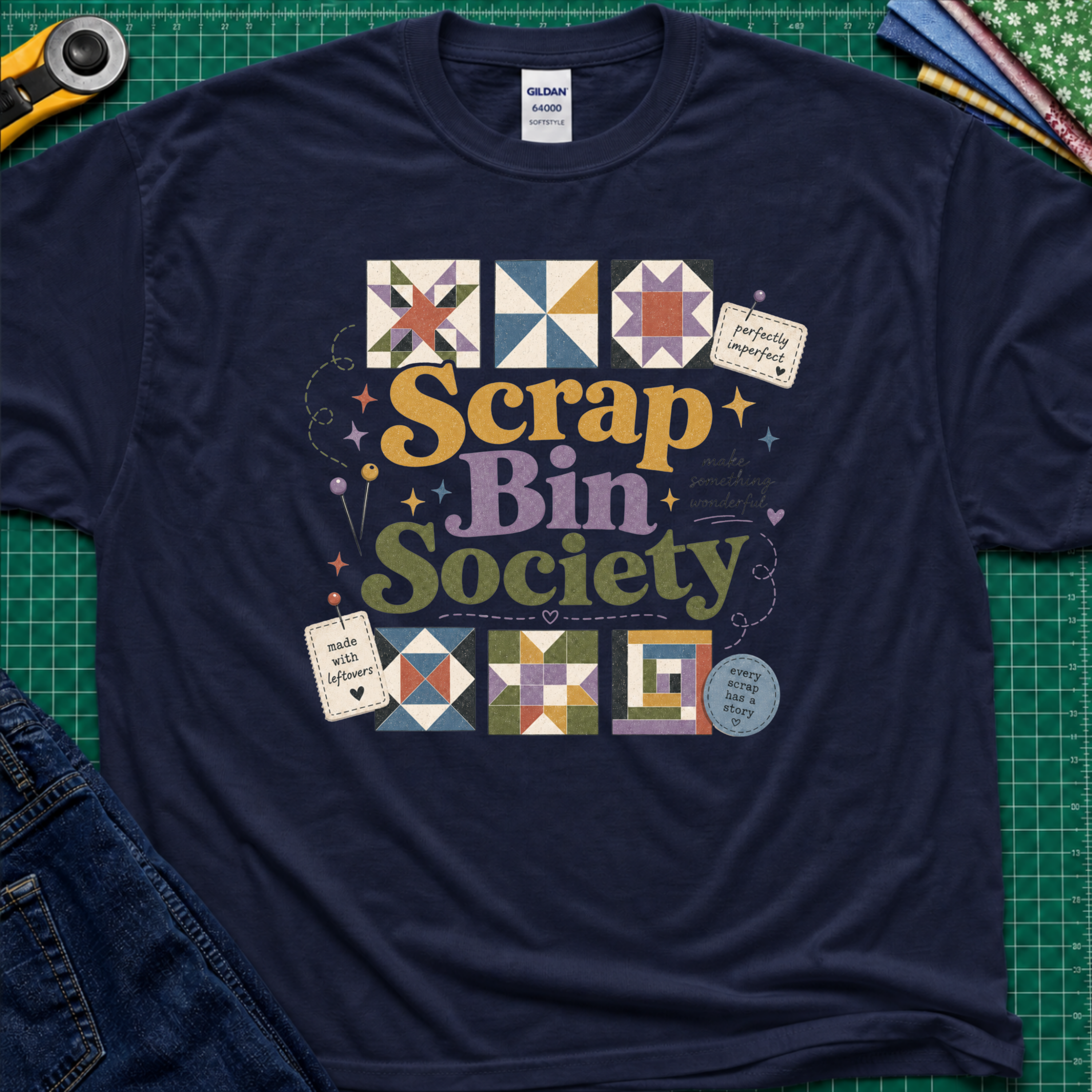 A navy blue Gildan t-shirt with a colorful 'Scrap Bin Society' graphic, laid flat on a green cutting mat with craft tools and jeans visible around the edges.