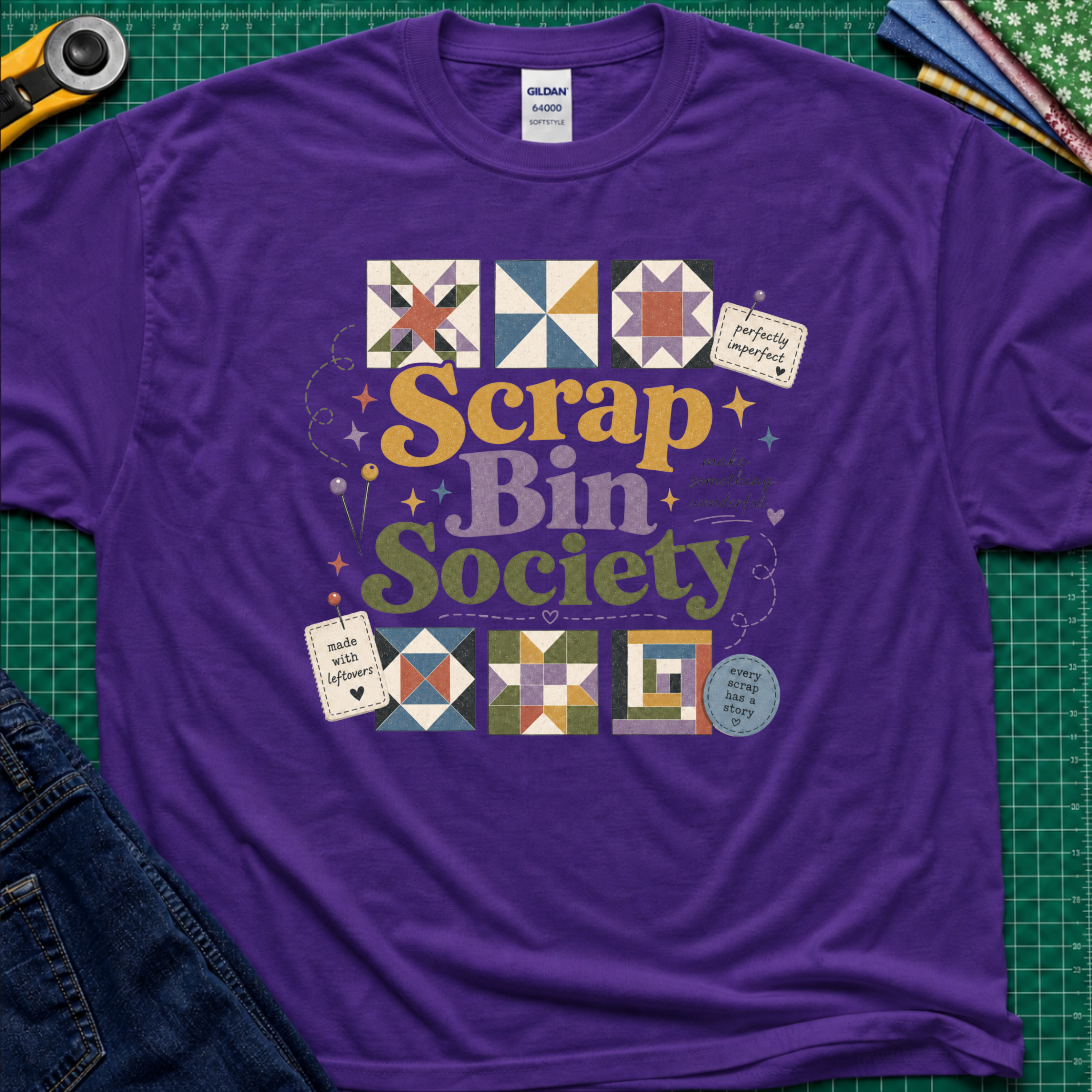 A purple Gildan t-shirt with a colorful 'Scrap Bin Society' graphic, laid flat on a green cutting mat with craft tools and jeans partially visible.