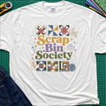 A white Gildan t-shirt featuring a colorful 'Scrap Bin Society' graphic with quilt patterns, laid flat on a green cutting mat with sewing tools and jeans partially visible.
