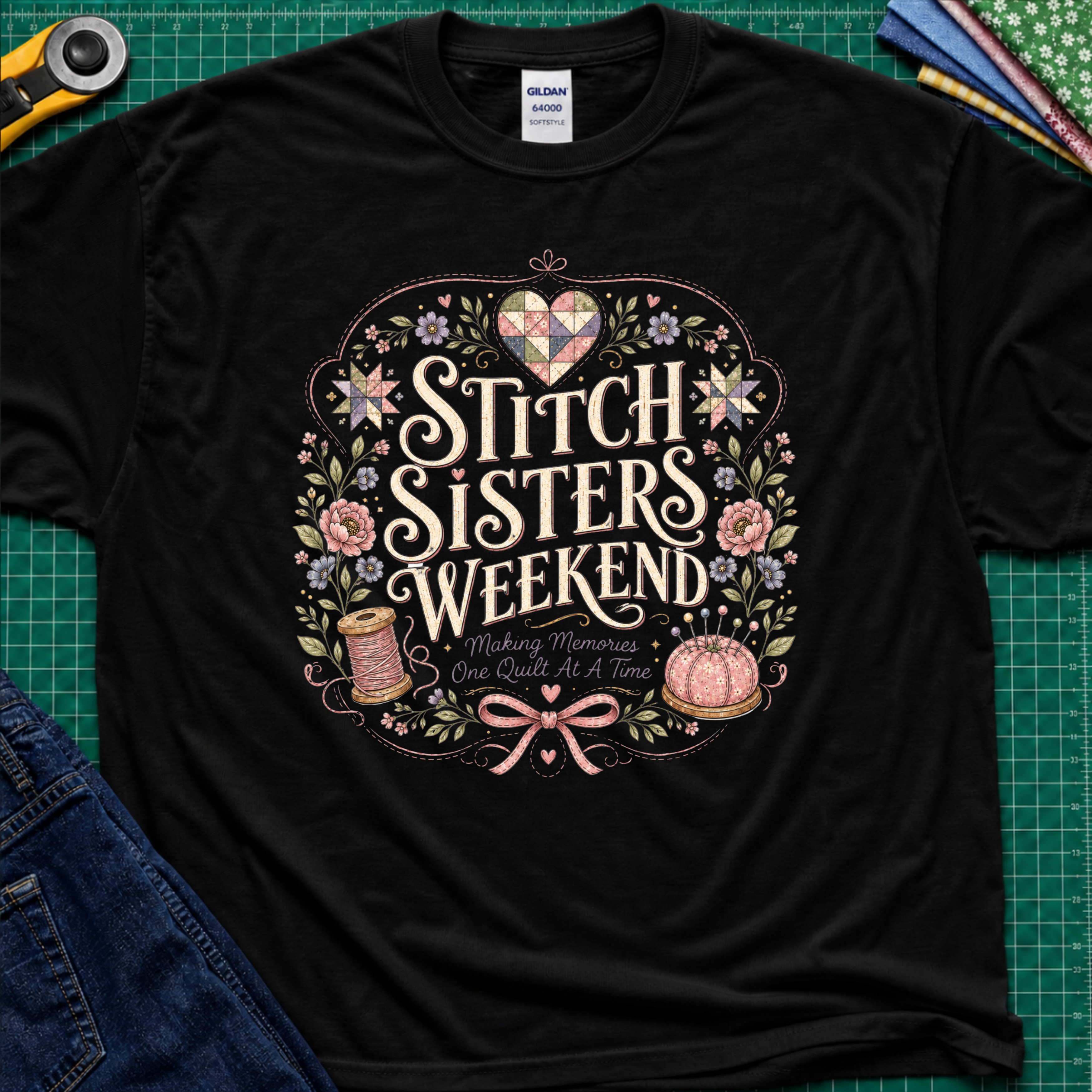 A black Gildan t-shirt featuring a colorful graphic that reads 'Stitch Sisters Weekend' surrounded by flowers, a spool of thread, pincushio and patchwork heart and stars. The shirt lays flat on a green cutting mat with rotary cutter, fabric stack and blue jeans partially visible in the corners.