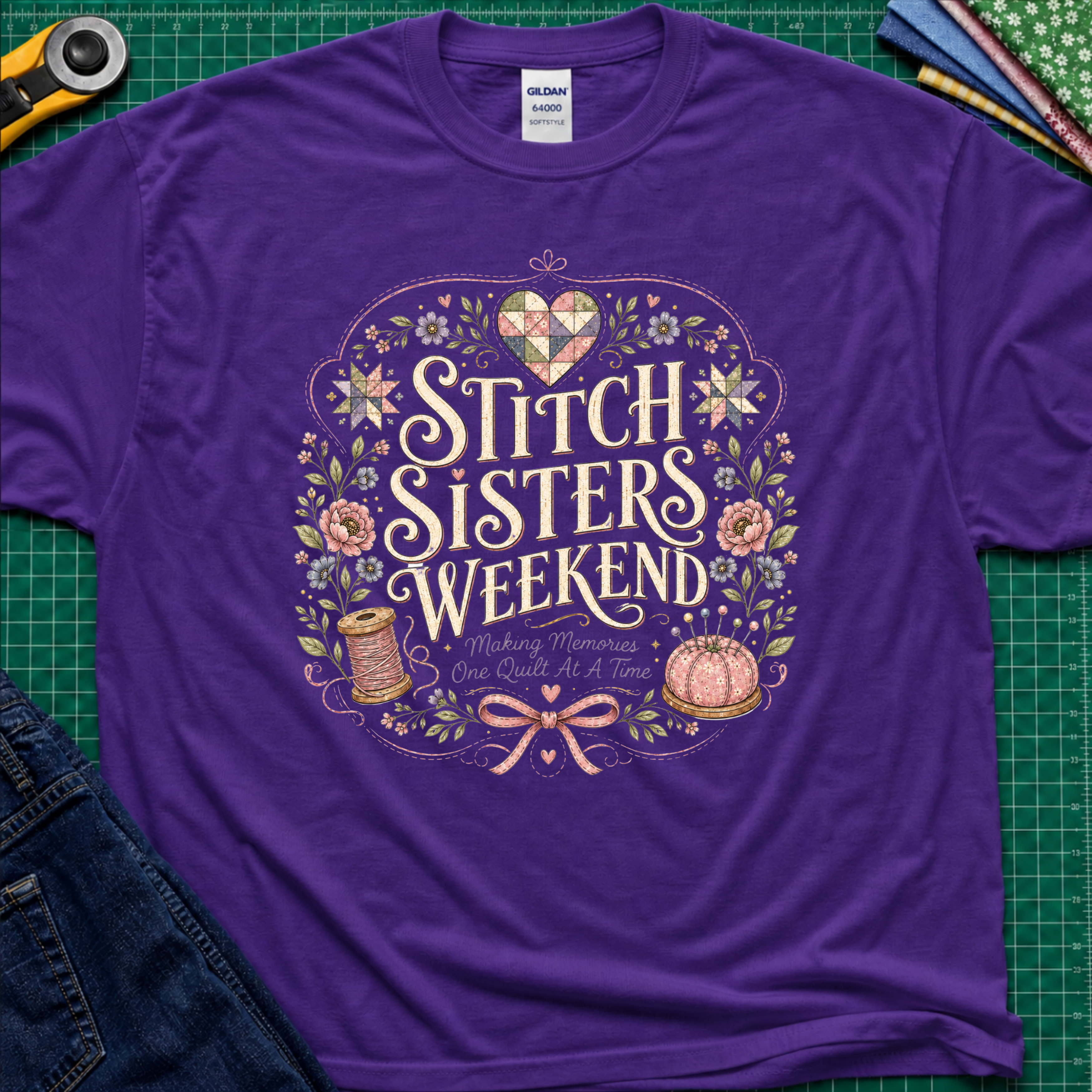 A purple Gildan t-shirt featuring a colorful graphic that reads 'Stitch Sisters Weekend' surrounded by flowers, a spool of thread, pincushio and patchwork heart and stars. The shirt lays flat on a green cutting mat with rotary cutter, fabric stack and blue jeans partially visible in the corners.