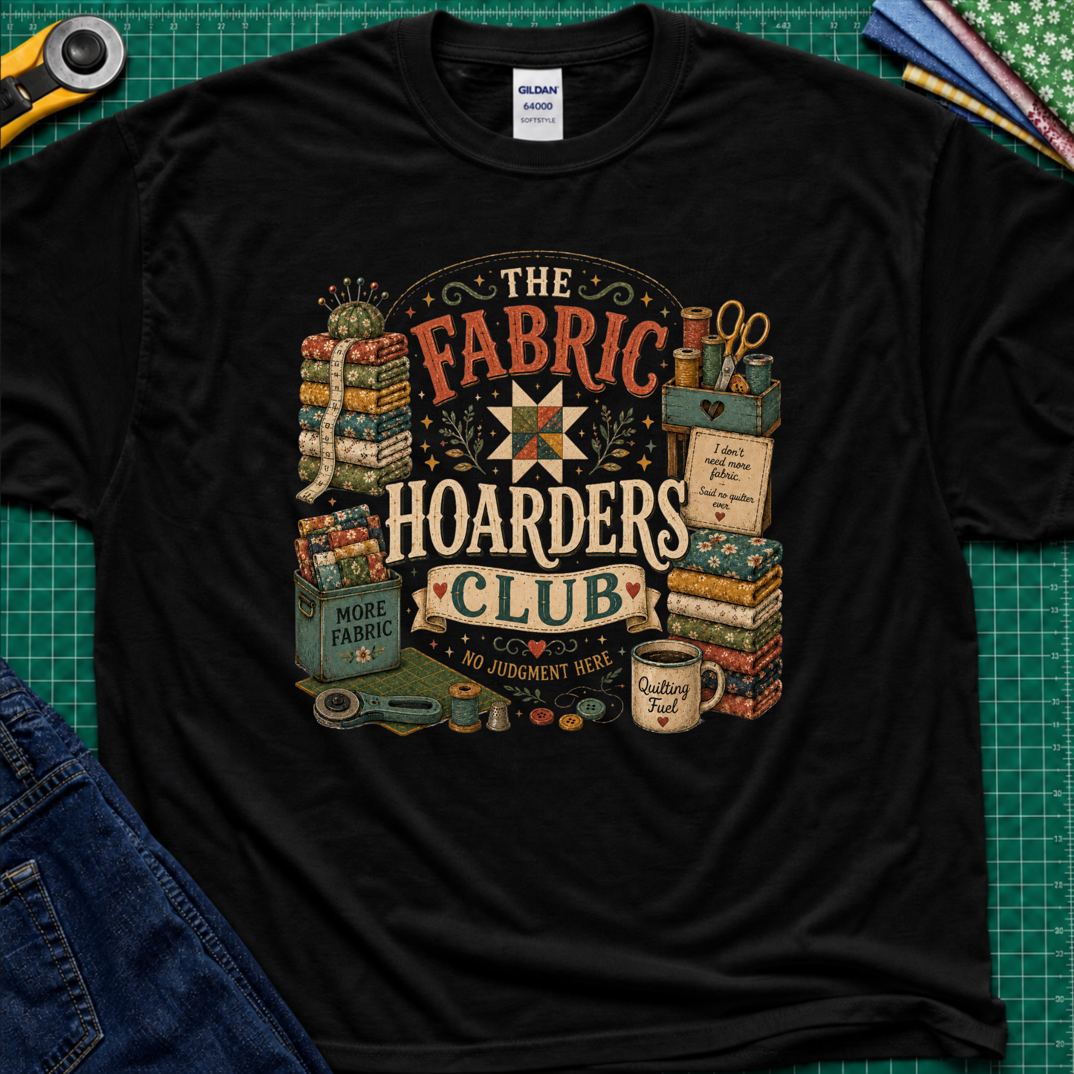 A black Gildan t-shirt laid flat on a green cutting mat, featuring a colorful vintage-style graphic that reads 'The Fabric Hoarders Club' with quilting supplies, fabric stacks, scissors, and a mug. Partial jeans and sewing tools are visible around the shirt.
