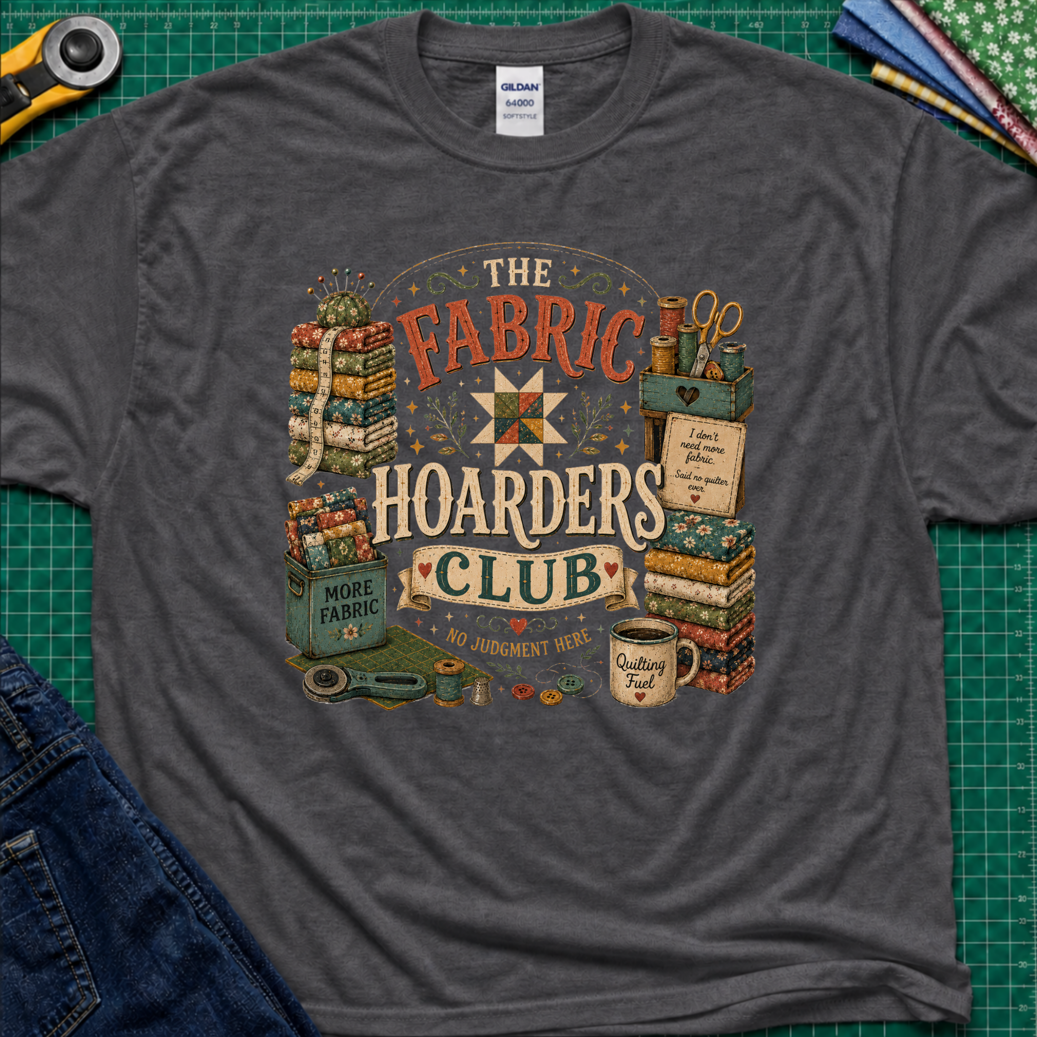 A dark grey Gildan t-shirt laid flat on a green cutting mat, featuring a colorful vintage-style graphic that reads 'The Fabric Hoarders Club' with quilting supplies, fabric stacks, scissors, and a mug. Partial jeans and sewing tools are visible around the shirt.