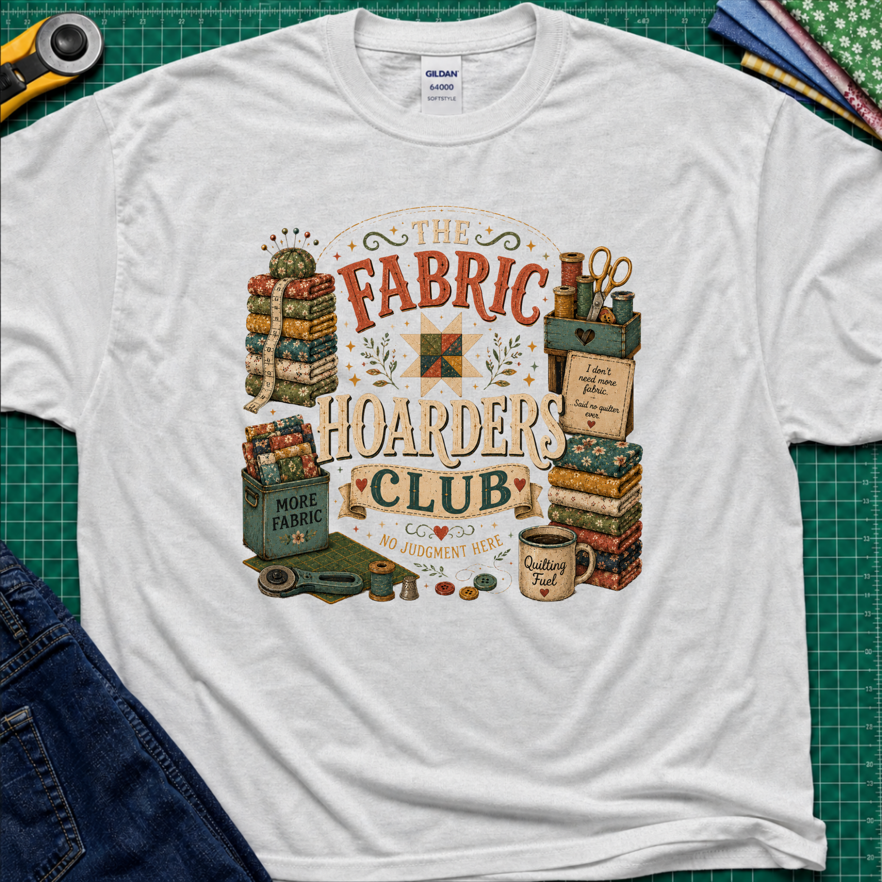 A light grey Gildan t-shirt laid flat on a green cutting mat, featuring a colorful vintage-style graphic that reads 'The Fabric Hoarders Club' with quilting supplies, fabric stacks, scissors, and a mug. Partial jeans and sewing tools are visible around the shirt.