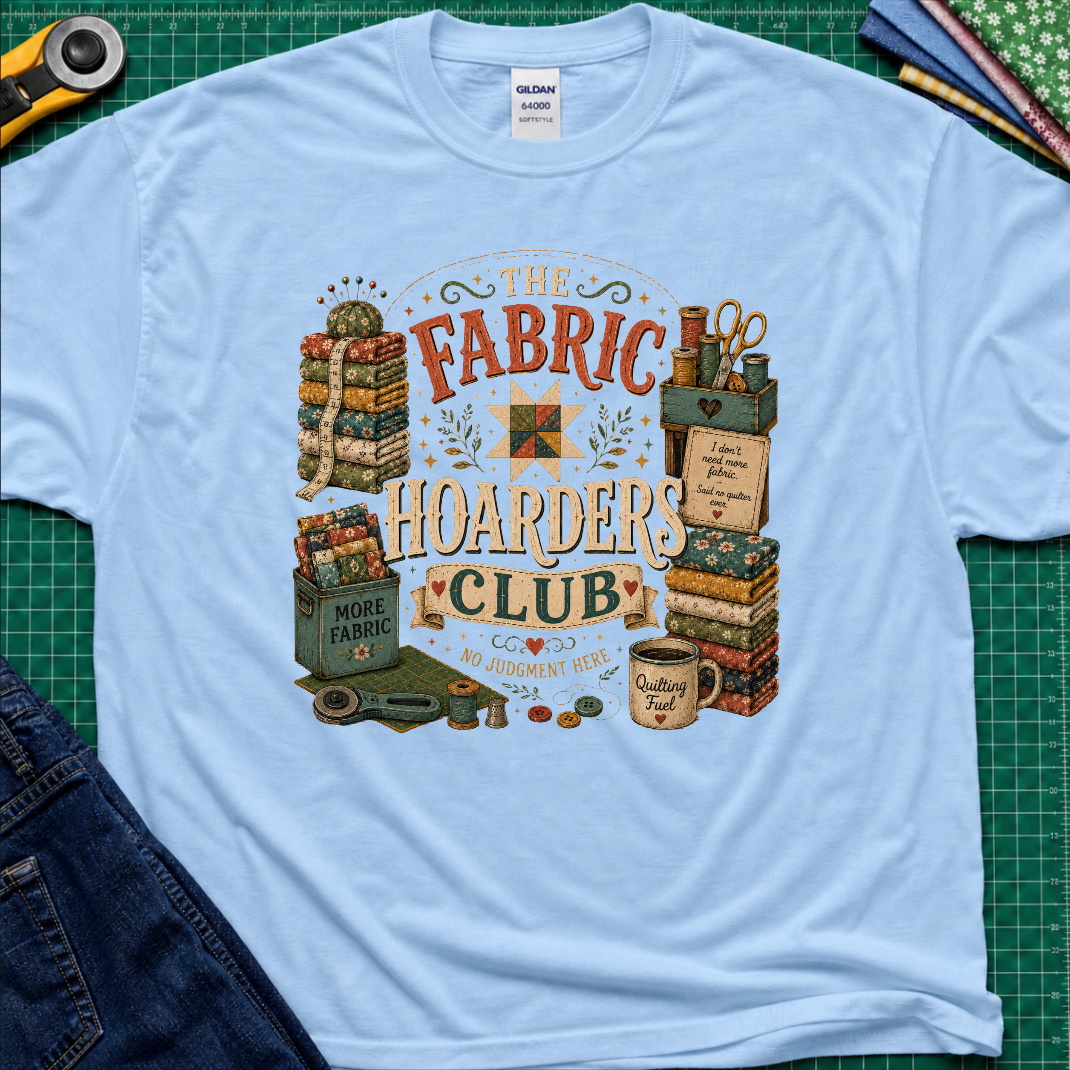 A light blue Gildan t-shirt laid flat on a green cutting mat, featuring a colorful vintage-style graphic that reads 'The Fabric Hoarders Club' with quilting supplies, fabric stacks, scissors, and a mug. Partial jeans and sewing tools are visible around the shirt.