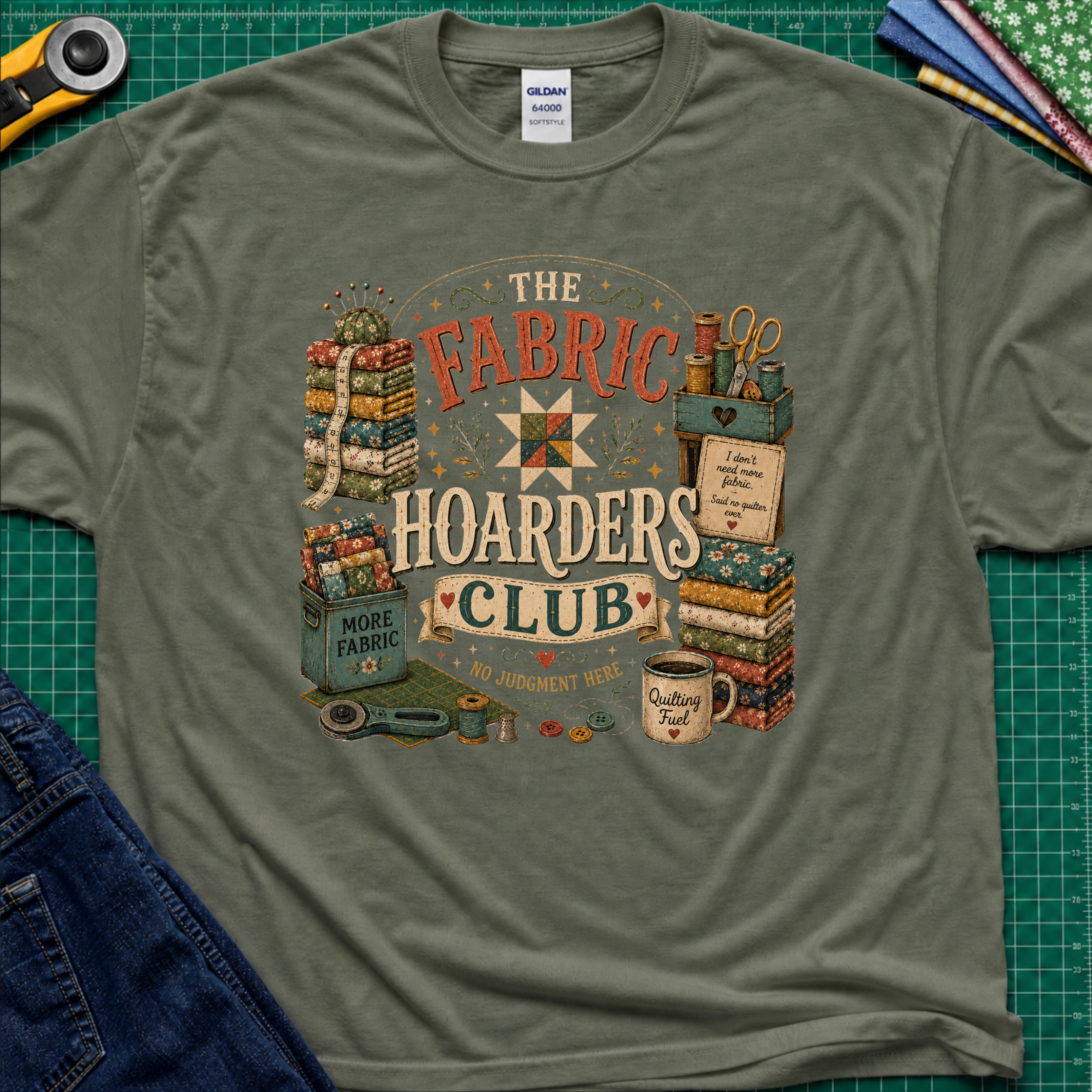 A military green Gildan t-shirt laid flat on a green cutting mat, featuring a colorful vintage-style graphic that reads 'The Fabric Hoarders Club' with quilting supplies, fabric stacks, scissors, and a mug. Partial jeans and sewing tools are visible around the shirt.