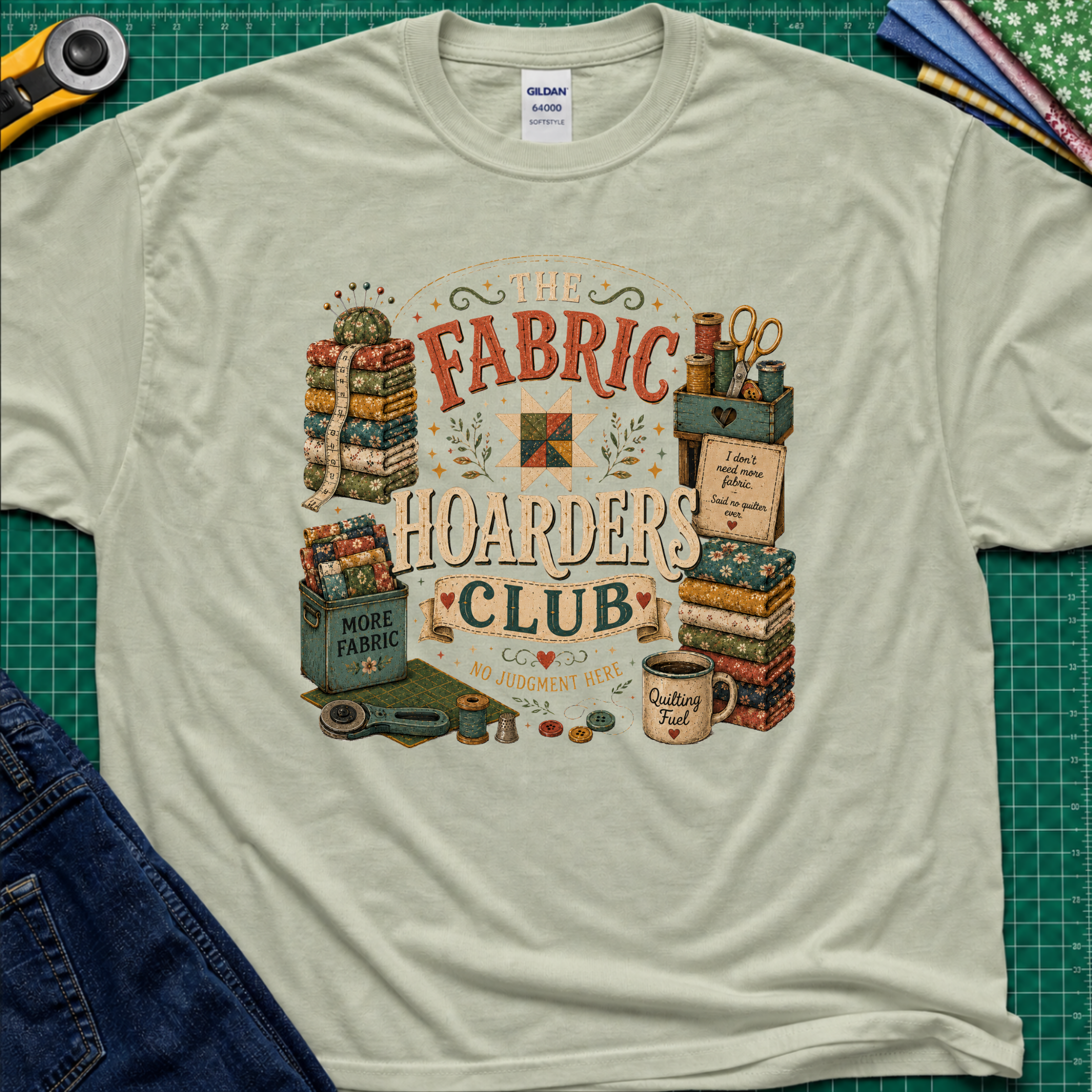 A light sage green Gildan t-shirt laid flat on a green cutting mat, featuring a colorful vintage-style graphic that reads 'The Fabric Hoarders Club' with quilting supplies, fabric stacks, scissors, and a mug. Partial jeans and sewing tools are visible around the shirt.