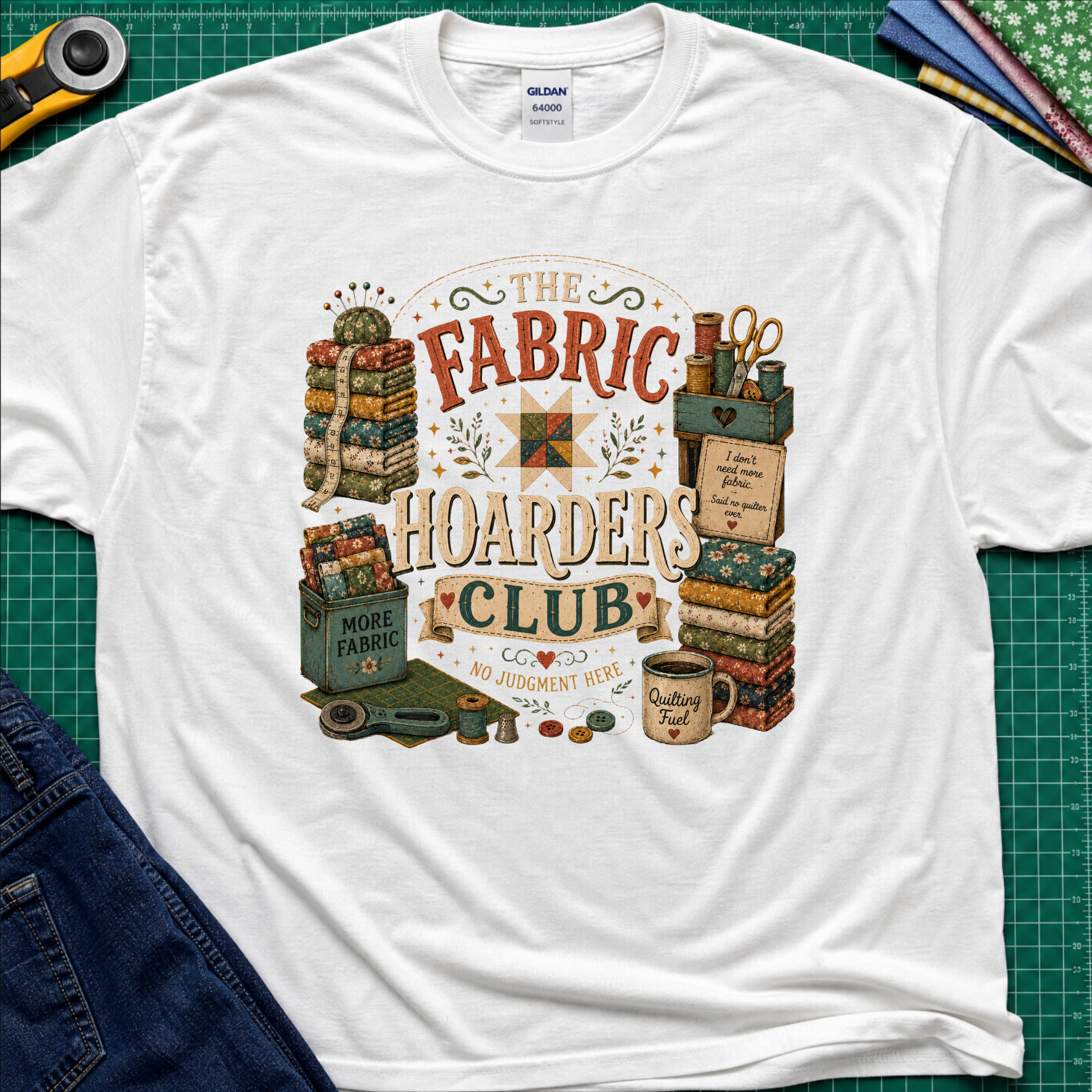 A white Gildan t-shirt laid flat on a green cutting mat, featuring a colorful vintage-style graphic that reads 'The Fabric Hoarders Club' with quilting supplies, fabric stacks, scissors, and a mug. Partial jeans and sewing tools are visible around the shirt.