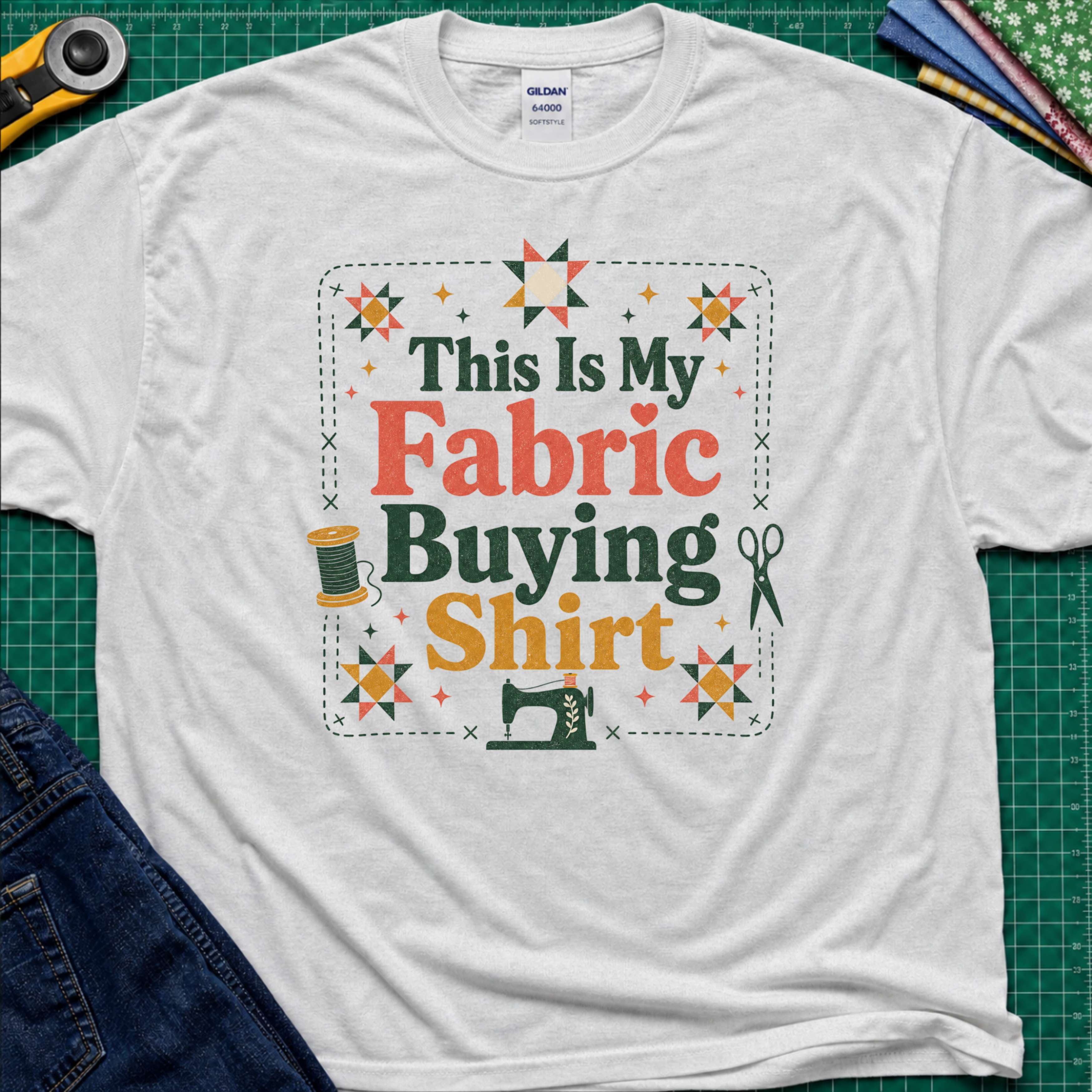 An light grey Gildan t-shirt with a colorful graphic that reads 'This Is My Fabric Buying Shirt' surrounded by sewing-themed illustrations like stars, a spool, scissors, and a sewing machine. The shirt is laid flat on a green cutting mat with sewing props around it.