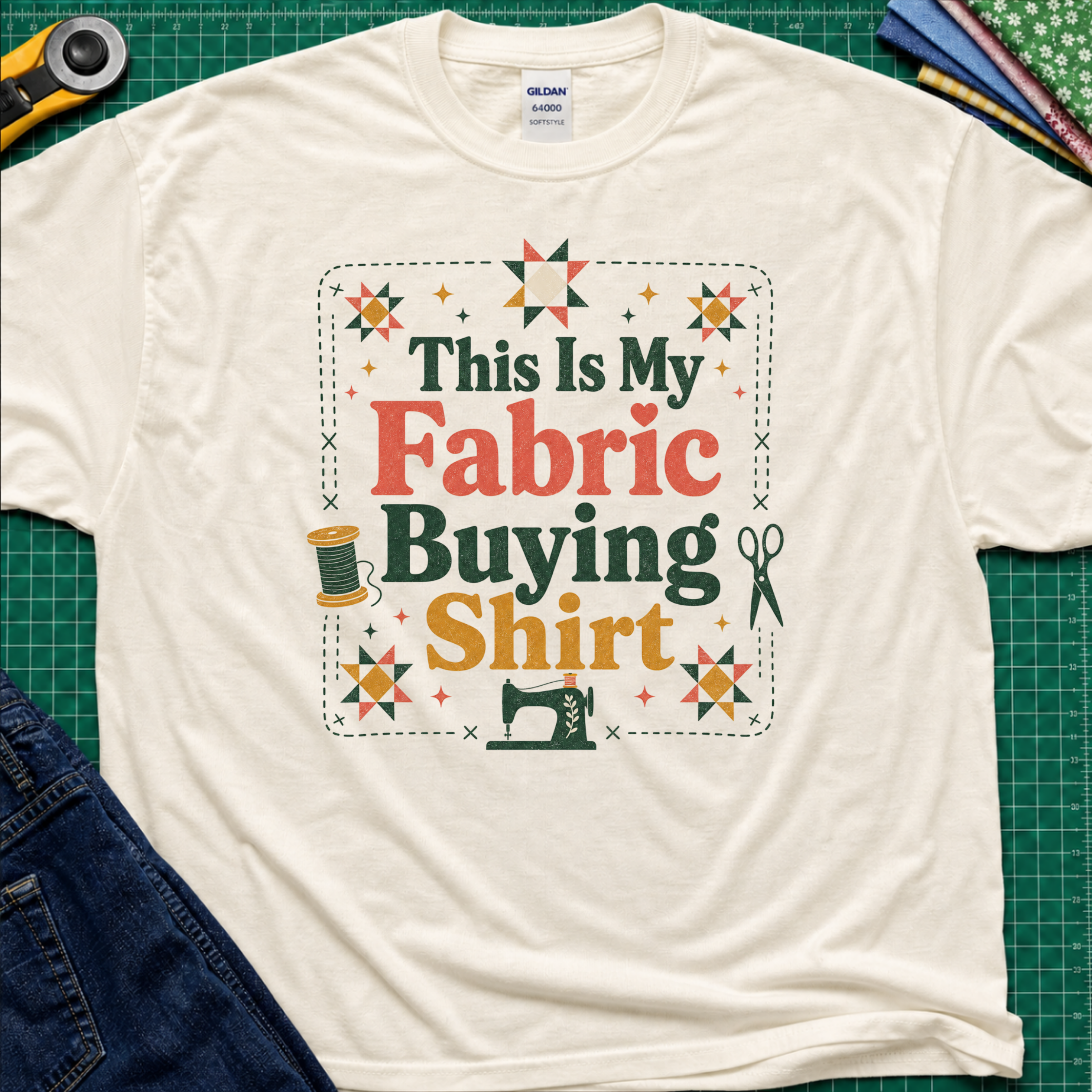 An light cream Gildan t-shirt with a colorful graphic that reads 'This Is My Fabric Buying Shirt' surrounded by sewing-themed illustrations like stars, a spool, scissors, and a sewing machine. The shirt is laid flat on a green cutting mat with sewing props around it.