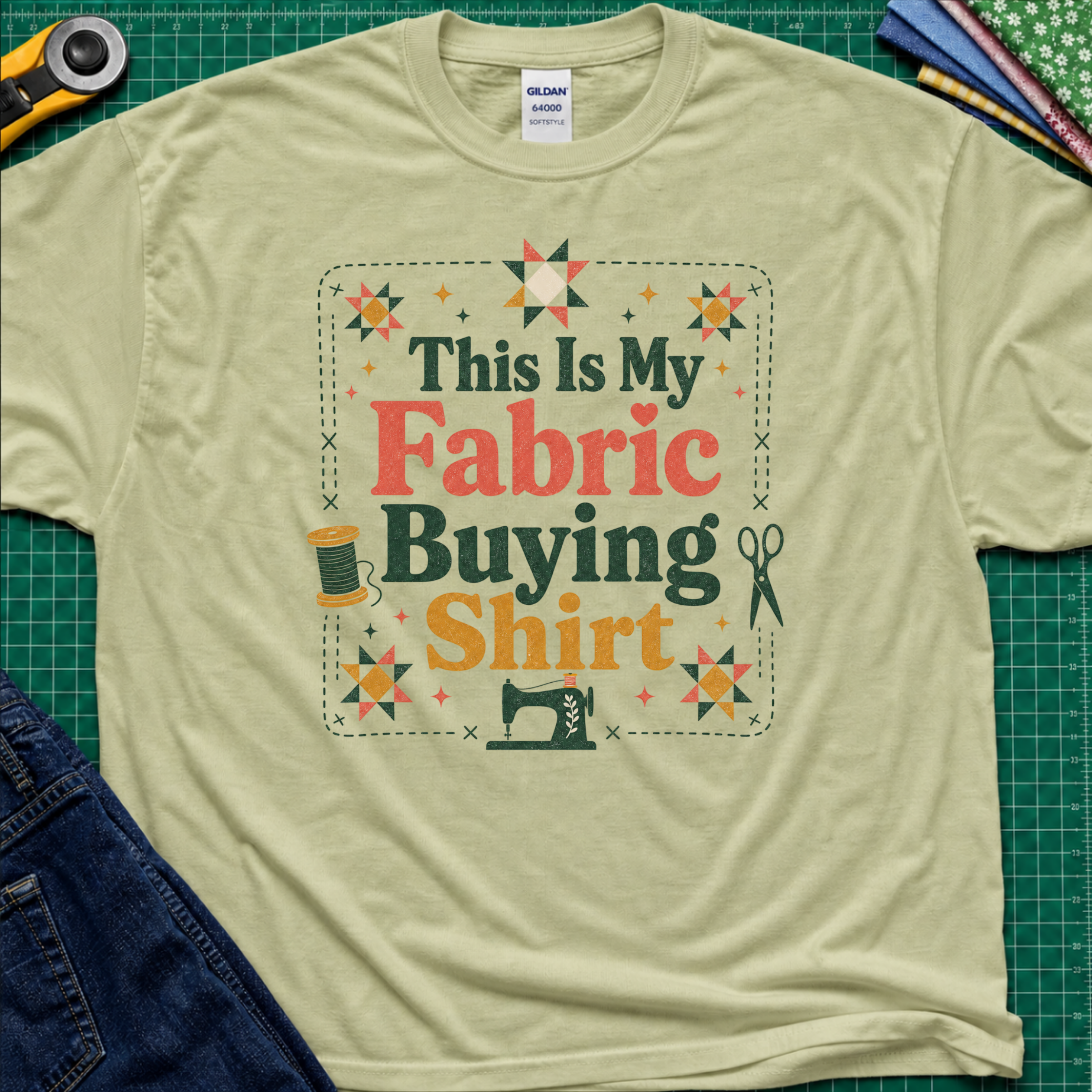 An light pistachio green Gildan t-shirt with a colorful graphic that reads 'This Is My Fabric Buying Shirt' surrounded by sewing-themed illustrations like stars, a spool, scissors, and a sewing machine. The shirt is laid flat on a green cutting mat with sewing props around it.