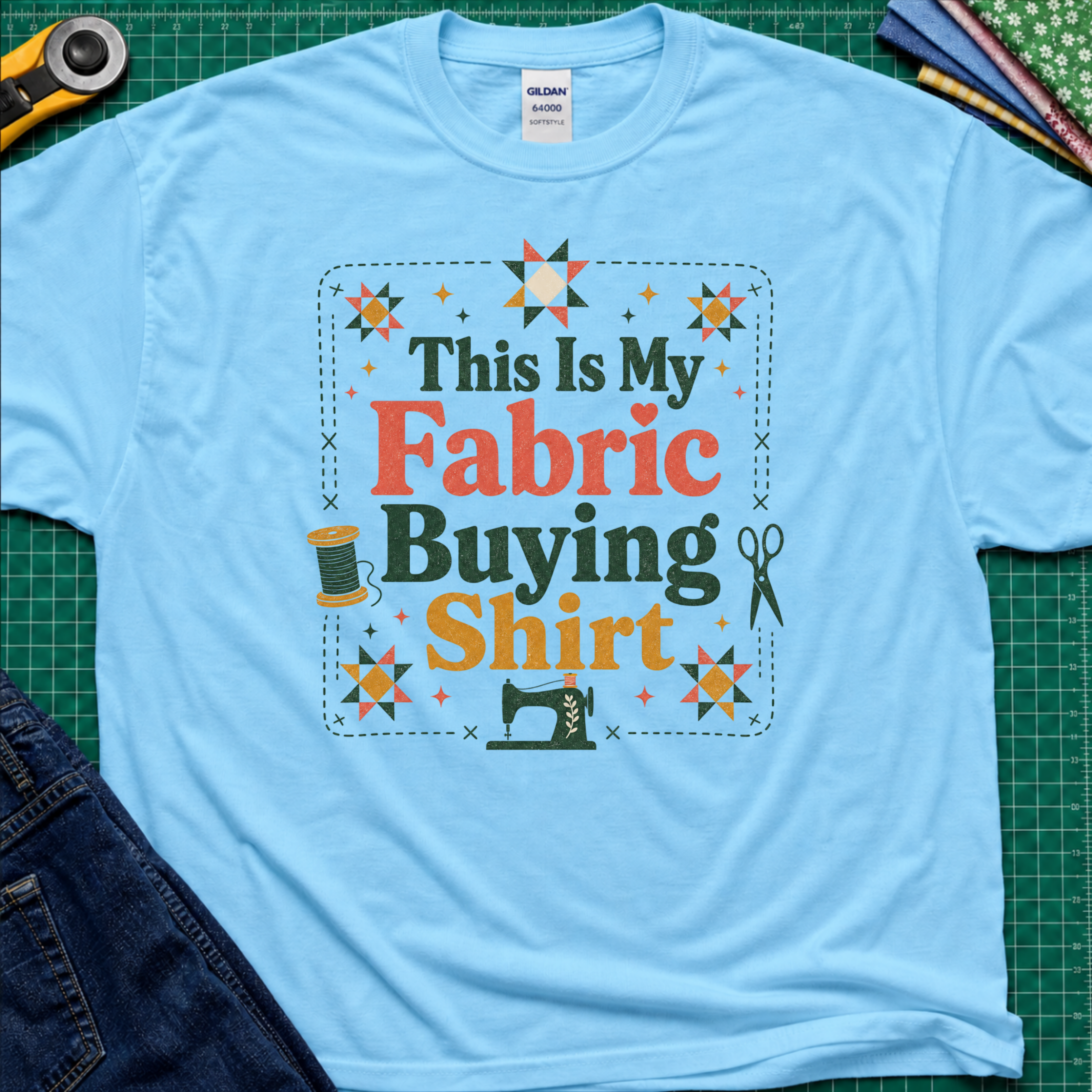 An light sky blue Gildan t-shirt with a colorful graphic that reads 'This Is My Fabric Buying Shirt' surrounded by sewing-themed illustrations like stars, a spool, scissors, and a sewing machine. The shirt is laid flat on a green cutting mat with sewing props around it.