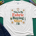 An white Gildan t-shirt with a colorful graphic that reads 'This Is My Fabric Buying Shirt' surrounded by sewing-themed illustrations like stars, a spool, scissors, and a sewing machine. The shirt is laid flat on a green cutting mat with sewing props around it.