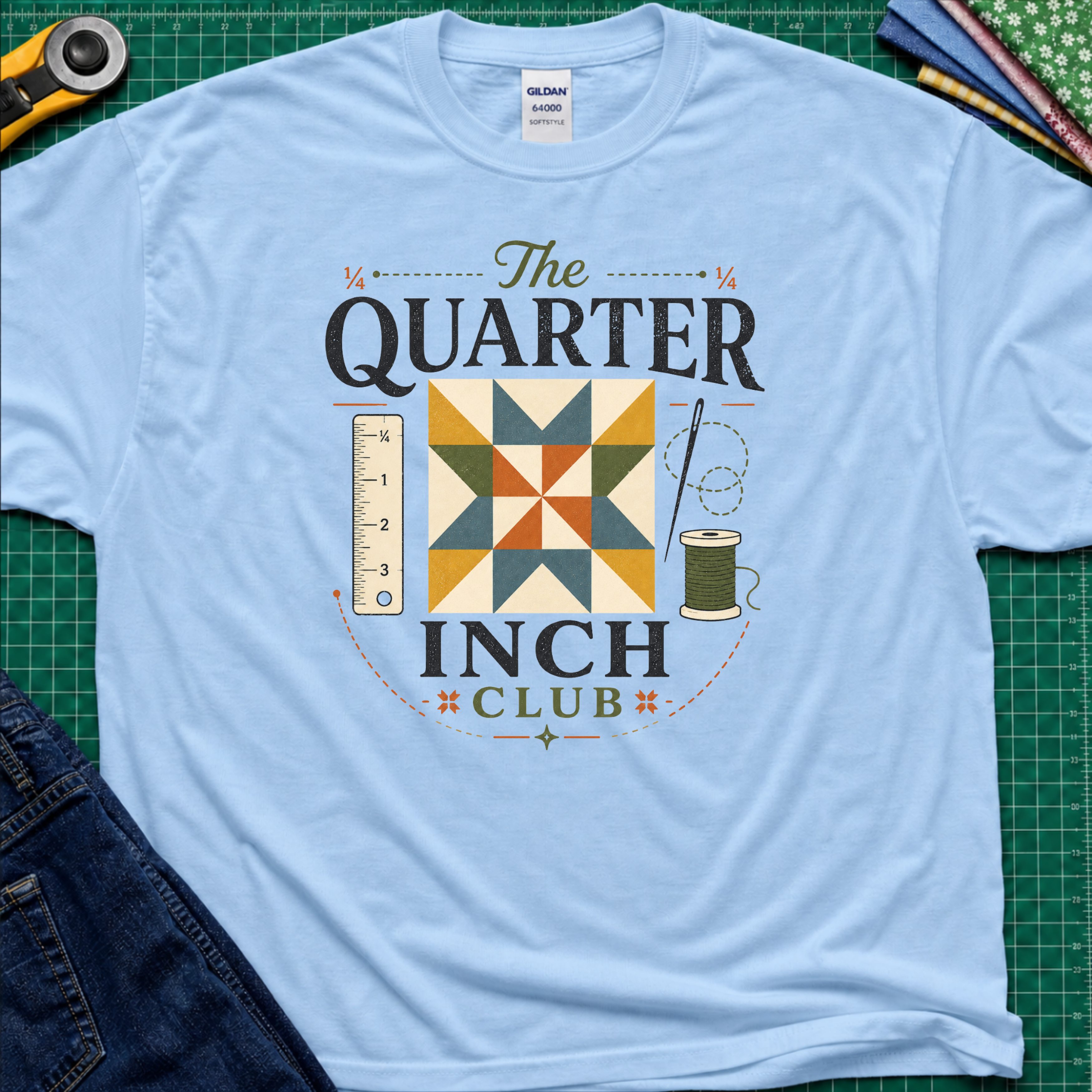 A light blue Gildan t-shirt with a retro-style quilted graphic that reads 'The QUARTER INCH CLUB'. The graphic features a geometric quilt pattern in yellow, orange, and blue, alongside a sewing ruler, spool, and needle.
