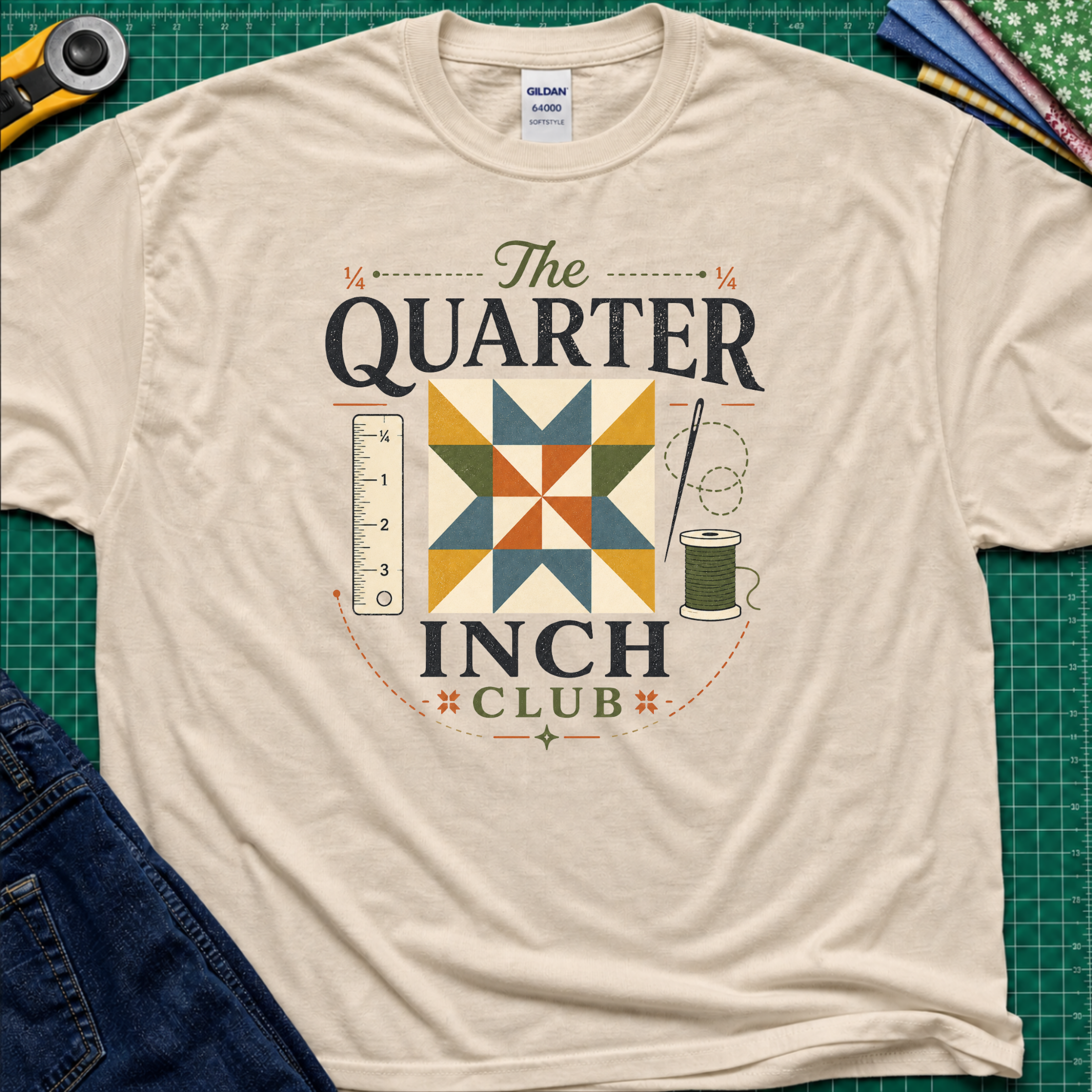 A dark cream Gildan t-shirt with a retro-style quilted graphic that reads 'The QUARTER INCH CLUB'. The graphic features a geometric quilt pattern in yellow, orange, and blue, alongside a sewing ruler, spool, and needle.