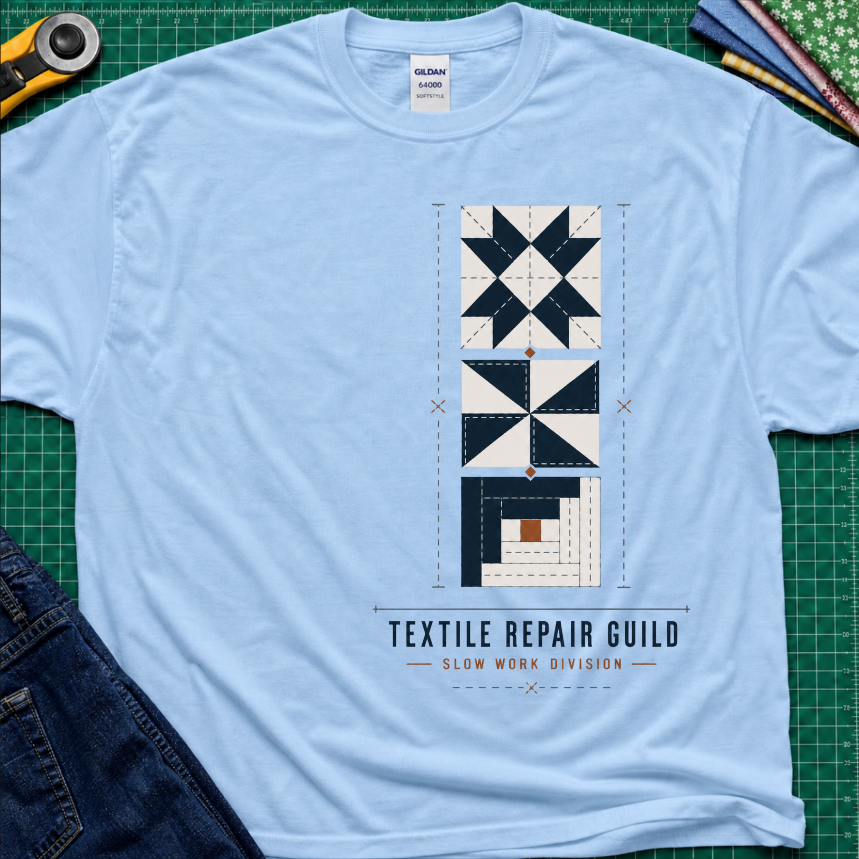 A light blue Gildan t-shirt featuring a graphic of geometric quilt blocks in navy blue, white and tan with the text 'TEXTILE REPAIR GUILD' and 'SLOW WORK DIVISION'. The shirt lays flat on a green cutting mat with a rotary cutter, fabric stack and blue jeans partially visible in the corners.