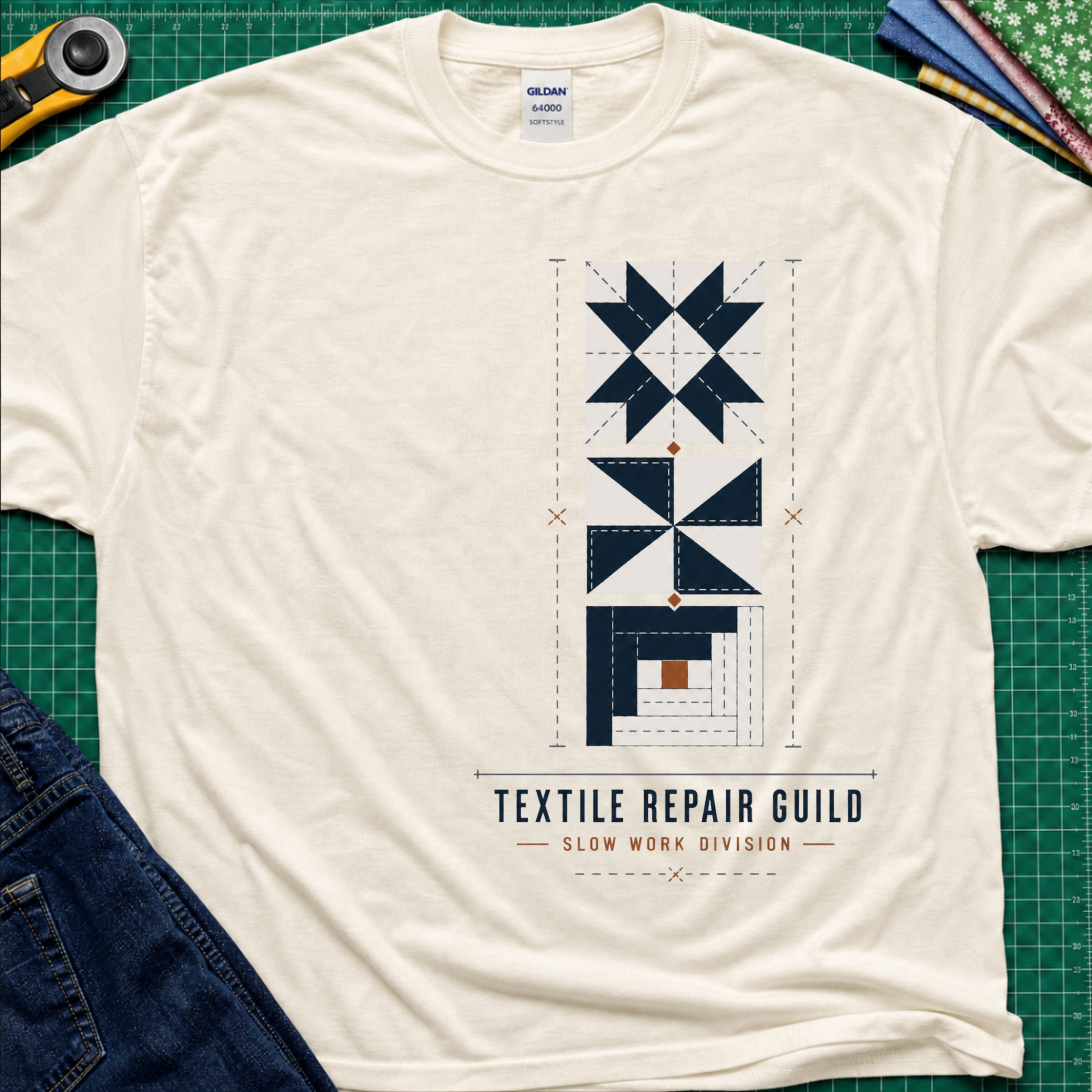 A light cream Gildan t-shirt featuring a graphic of geometric quilt blocks in navy blue, white and tan with the text 'TEXTILE REPAIR GUILD' and 'SLOW WORK DIVISION'. The shirt lays flat on a green cutting mat with a rotary cutter, fabric stack and blue jeans partially visible in the corners.