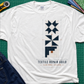 A white Gildan t-shirt featuring a graphic of geometric quilt blocks in navy blue, white and tan with the text 'TEXTILE REPAIR GUILD' and 'SLOW WORK DIVISION'. The shirt lays flat on a green cutting mat with a rotary cutter, fabric stack and blue jeans partially visible in the corners.