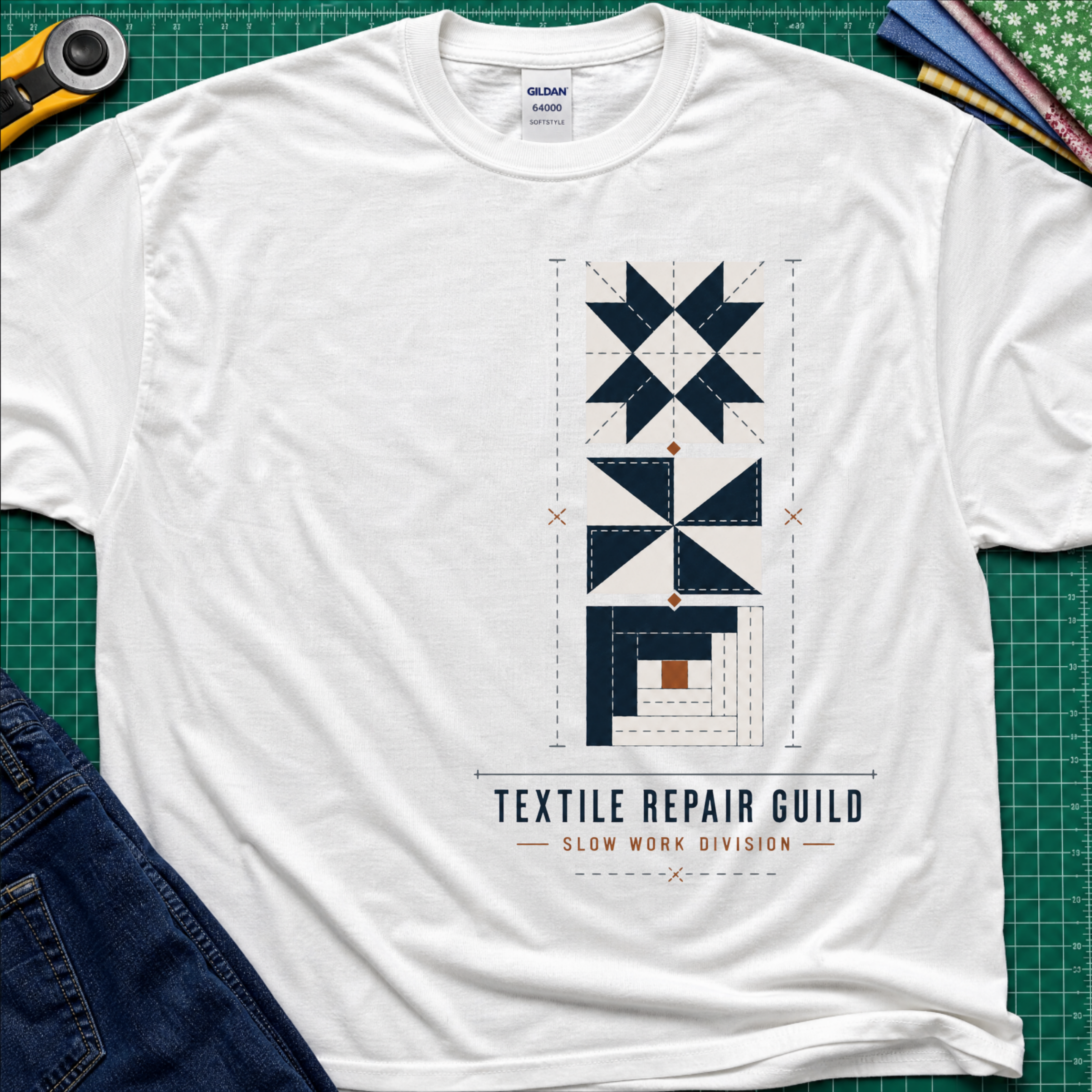 A white Gildan t-shirt featuring a graphic of geometric quilt blocks in navy blue, white and tan with the text 'TEXTILE REPAIR GUILD' and 'SLOW WORK DIVISION'. The shirt lays flat on a green cutting mat with a rotary cutter, fabric stack and blue jeans partially visible in the corners.