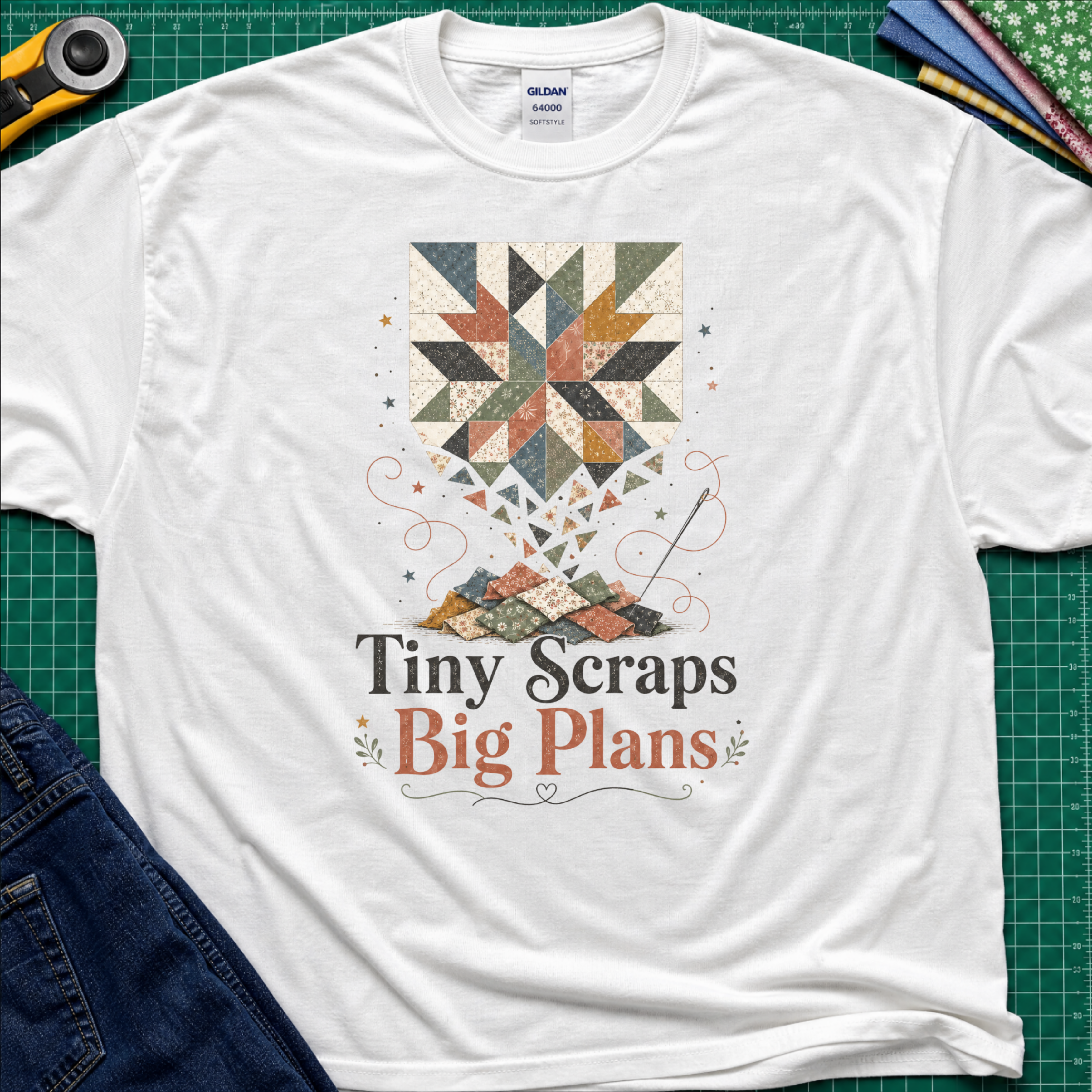 A white t-shirt laid flat on a green cutting mat background, featuring a quilt pattern graphic and the text 'Tiny Scraps Big Plans'. Crafting props like a rotary cutter, jeans, and fabric are partially visible around the shirt.