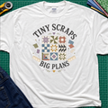 A white Gildan t-shirt laid flat on a green cutting mat background, featuring a colorful quilt pattern graphic with the text 'TINY SCRAPS BIG PLANS'. The shirt is styled with a rotary cutter, denim jeans, and folded fabric around it.