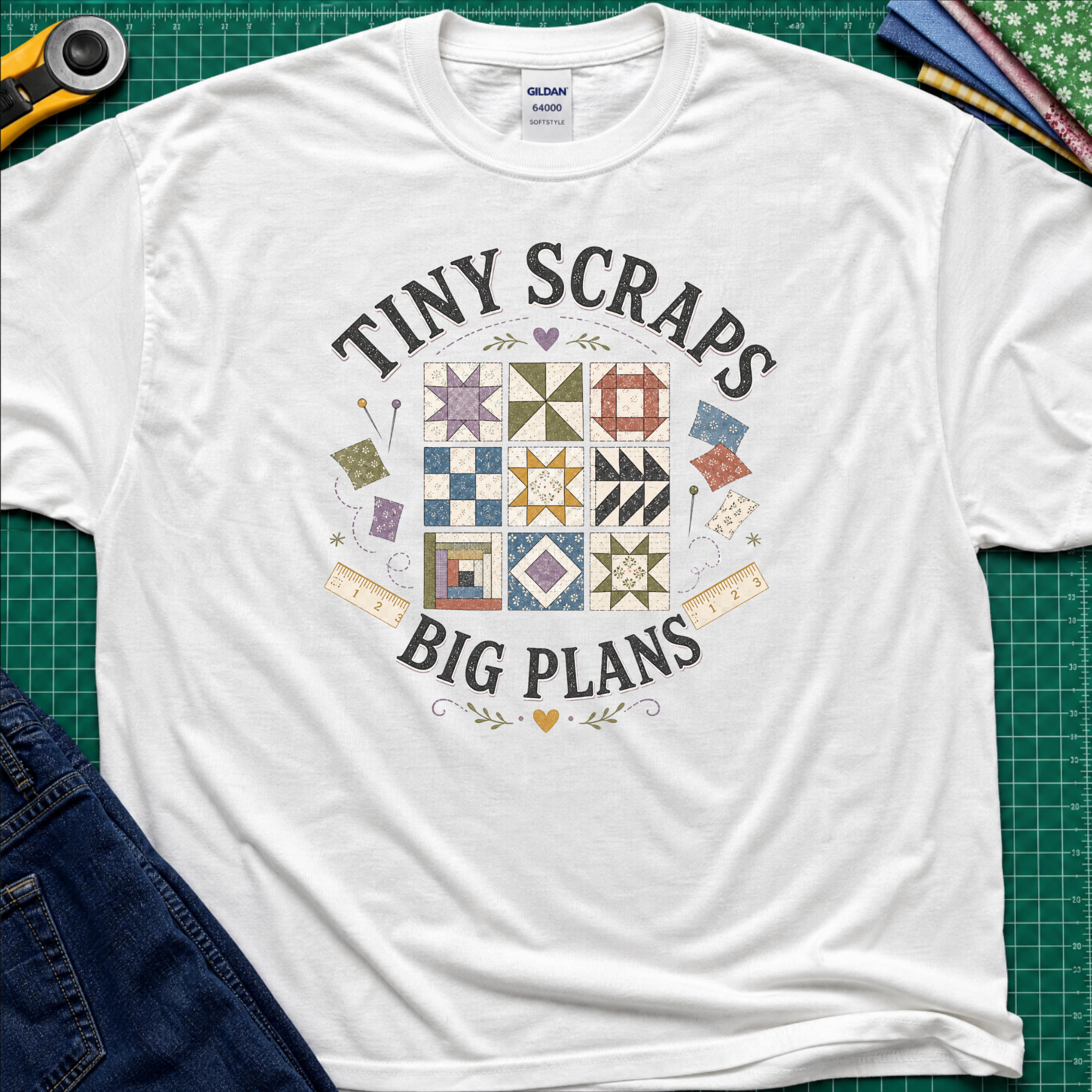 A white Gildan t-shirt laid flat on a green cutting mat background, featuring a colorful quilt pattern graphic with the text 'TINY SCRAPS BIG PLANS'. The shirt is styled with a rotary cutter, denim jeans, and folded fabric around it.
