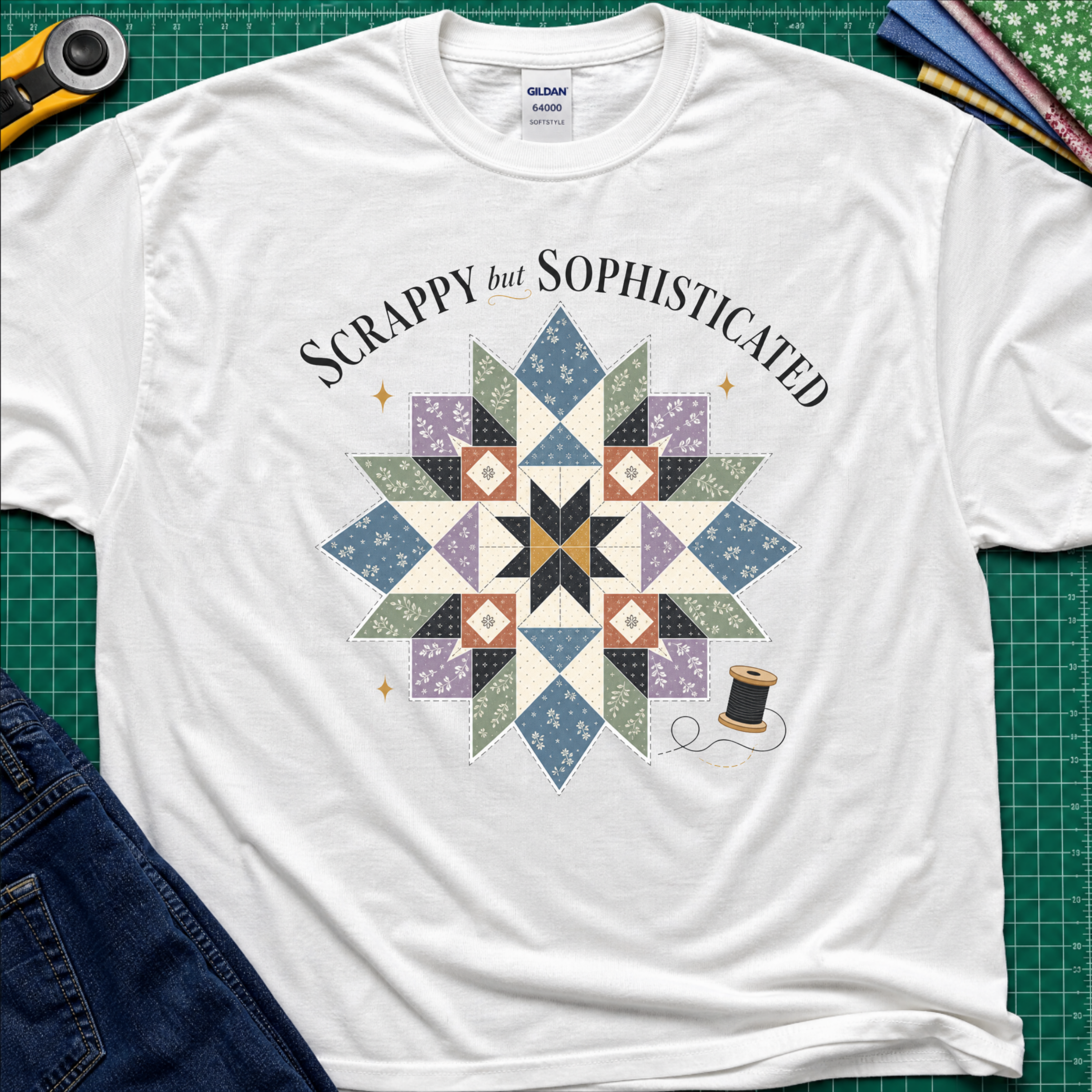 A black Gildan t-shirt with a colorful quilted starburst graphic and the text 'Scrappy but Sophisticated' printed on the front, styled on a green cutting mat with sewing tools and denim.