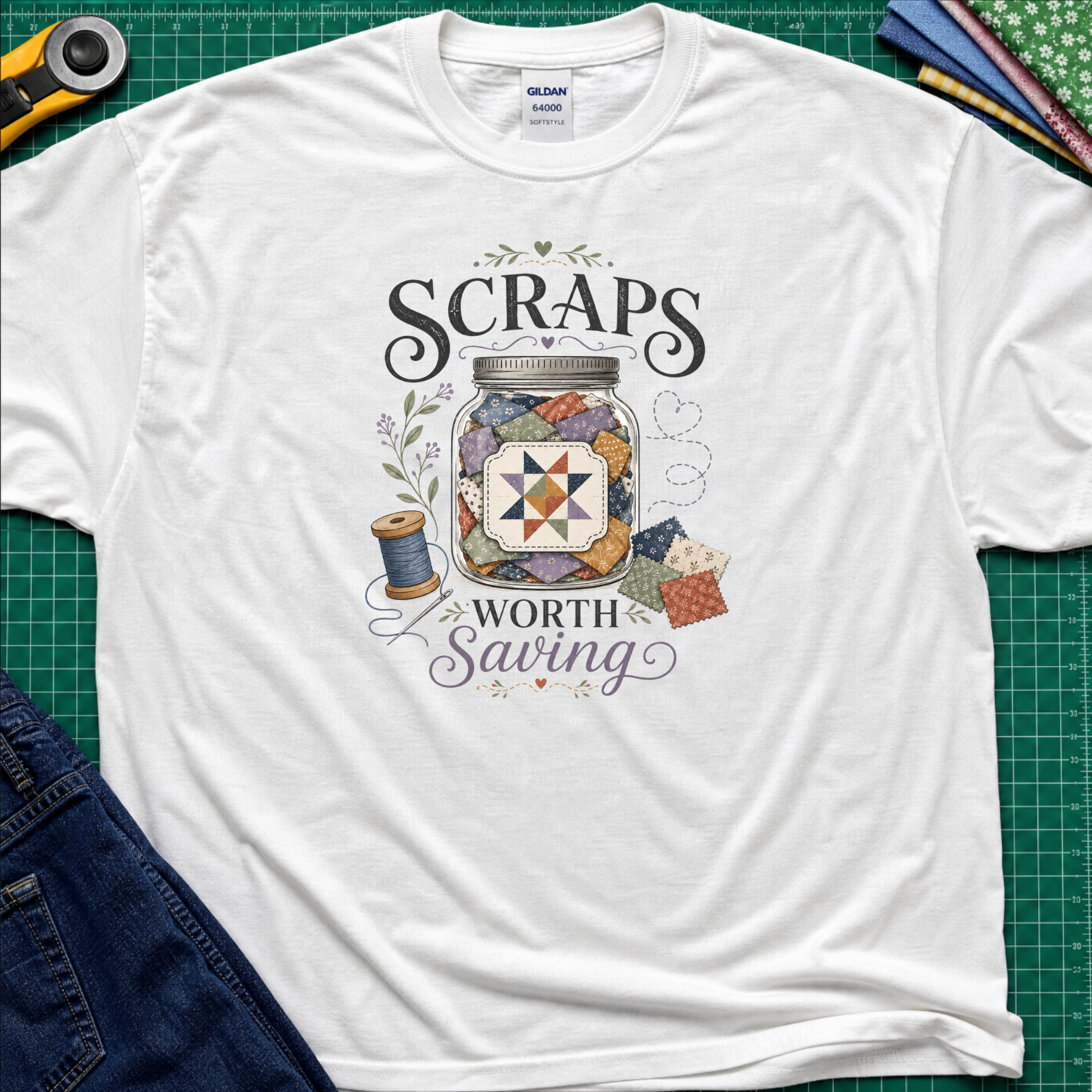 A white Gildan t-shirt laid flat on a green cutting mat, featuring a graphic design that reads 'SCRAPS WORTH Saving' around an illustration of a jar filled with scraps, thread spool, and fabric pieces. A rotary cutter, scissors, and folded fabrics are visible around the shirt as styling props.