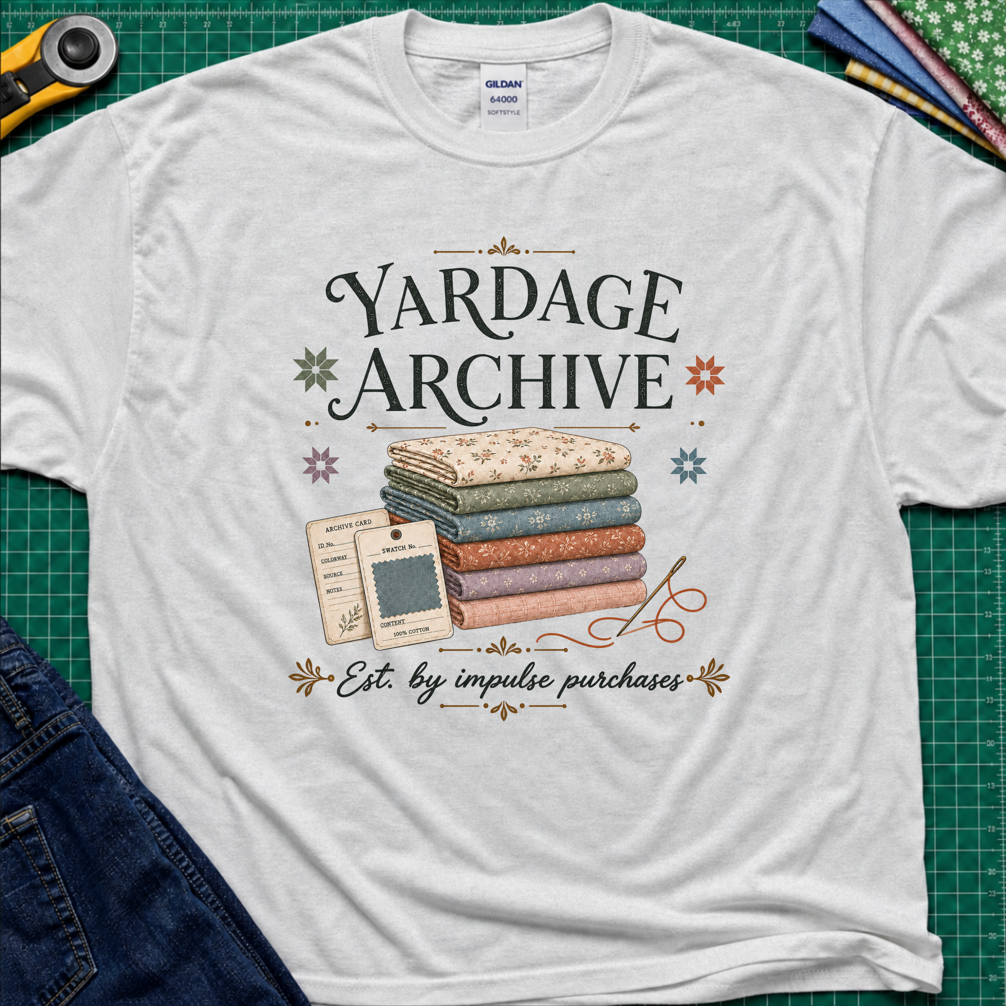 A light grey Gildan t-shirt laid flat on a green cutting mat, featuring a vintage-style graphic that reads 'Yardage Archive' with a stack of folded fabrics, a fabric label and swatch. Decorative craft tools and blue jeans are partially visible around the edges.