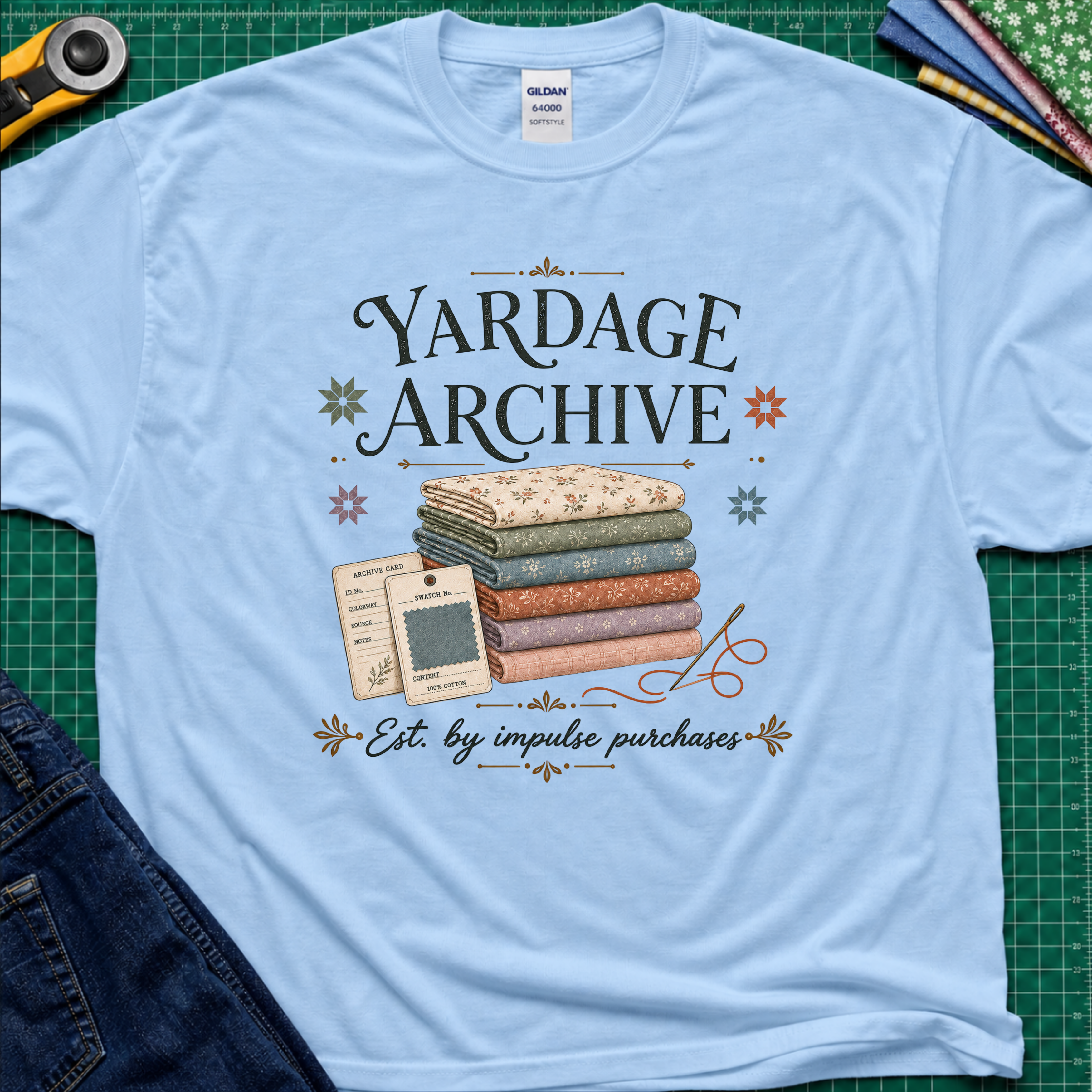 A light blue Gildan t-shirt laid flat on a green cutting mat, featuring a vintage-style graphic that reads 'Yardage Archive' with a stack of folded fabrics, a fabric label and swatch. Decorative craft tools and blue jeans are partially visible around the edges.