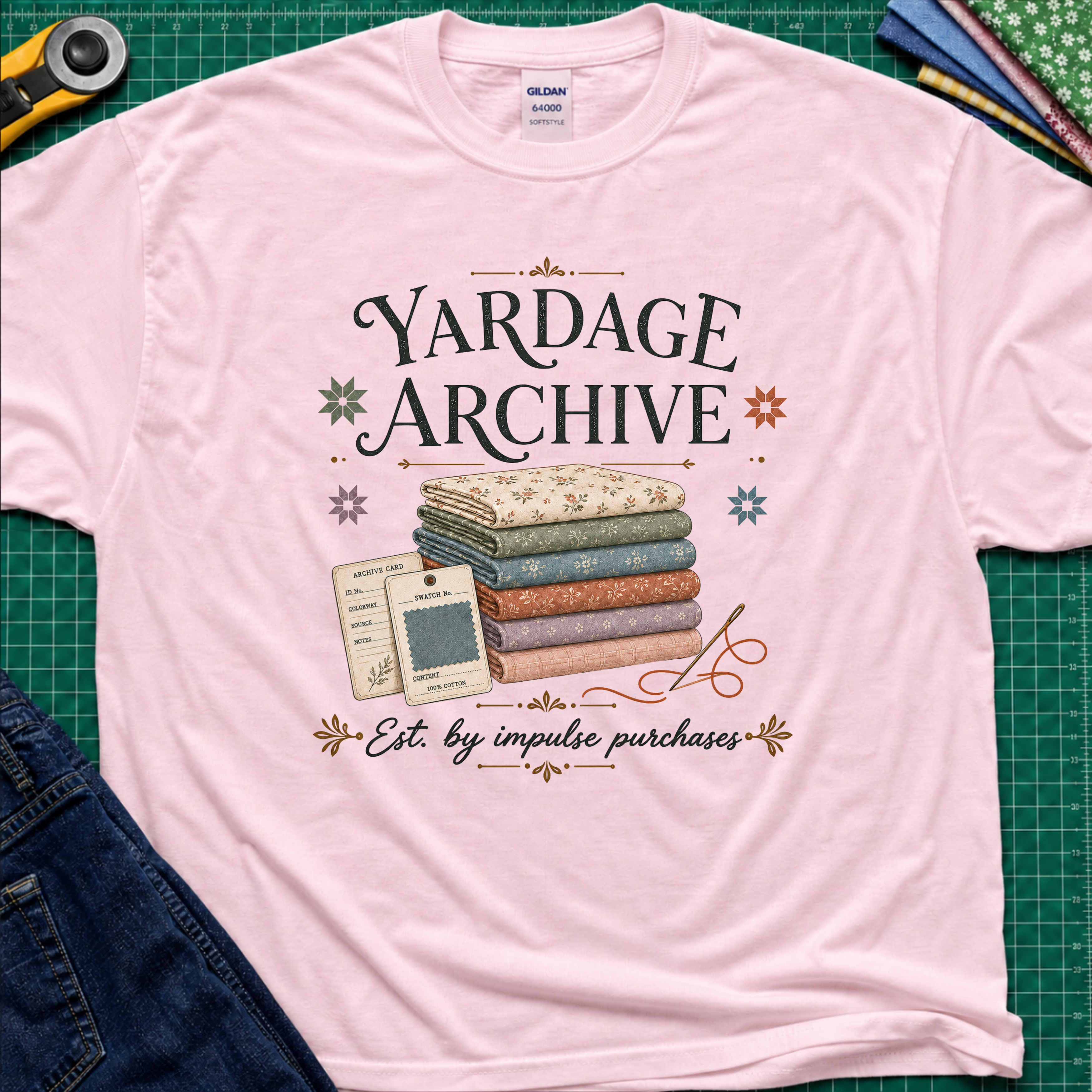 A light pink Gildan t-shirt laid flat on a green cutting mat, featuring a vintage-style graphic that reads 'Yardage Archive' with a stack of folded fabrics, a fabric label and swatch. Decorative craft tools and blue jeans are partially visible around the edges.