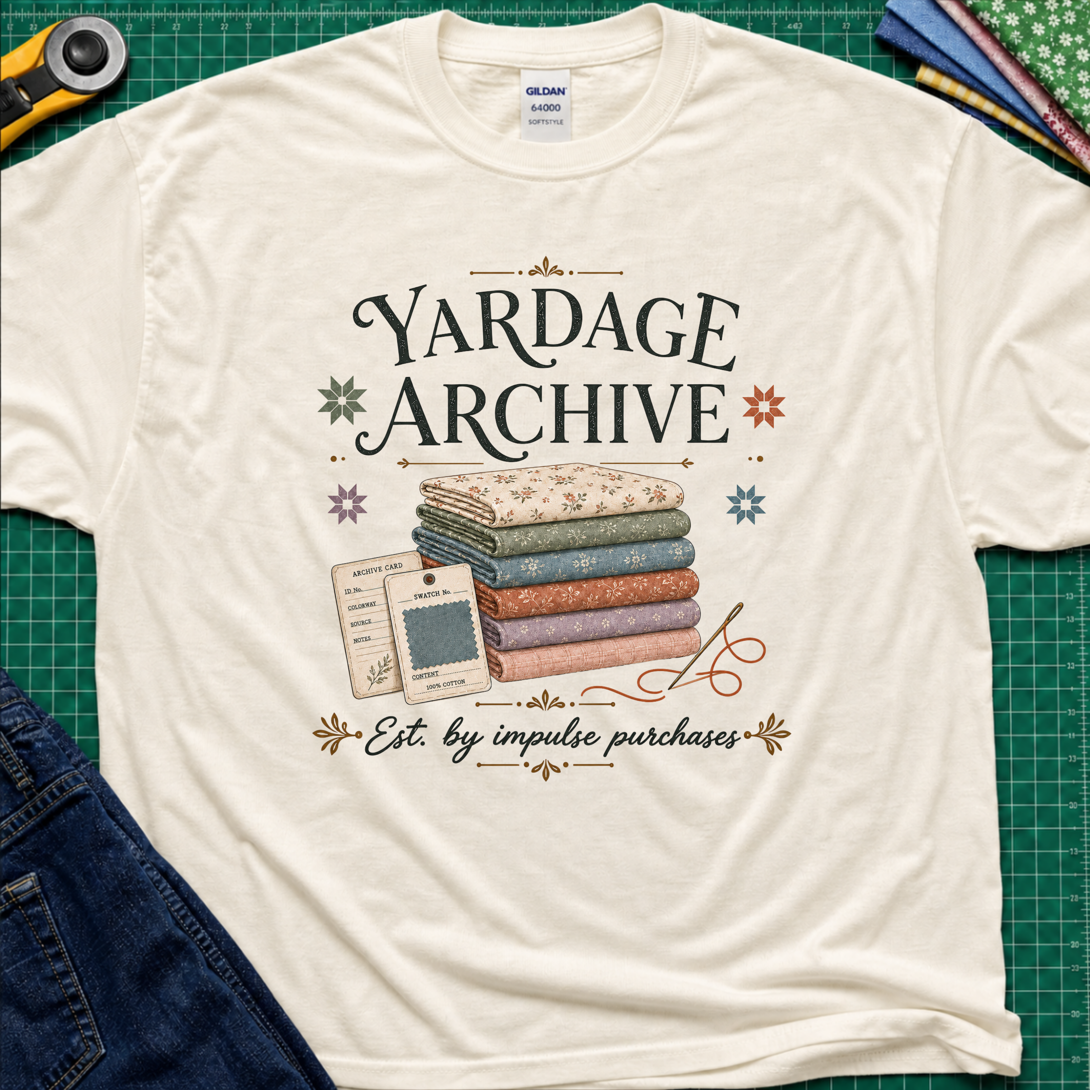 A light cream Gildan t-shirt laid flat on a green cutting mat, featuring a vintage-style graphic that reads 'Yardage Archive' with a stack of folded fabrics, a fabric label and swatch. Decorative craft tools and blue jeans are partially visible around the edges.