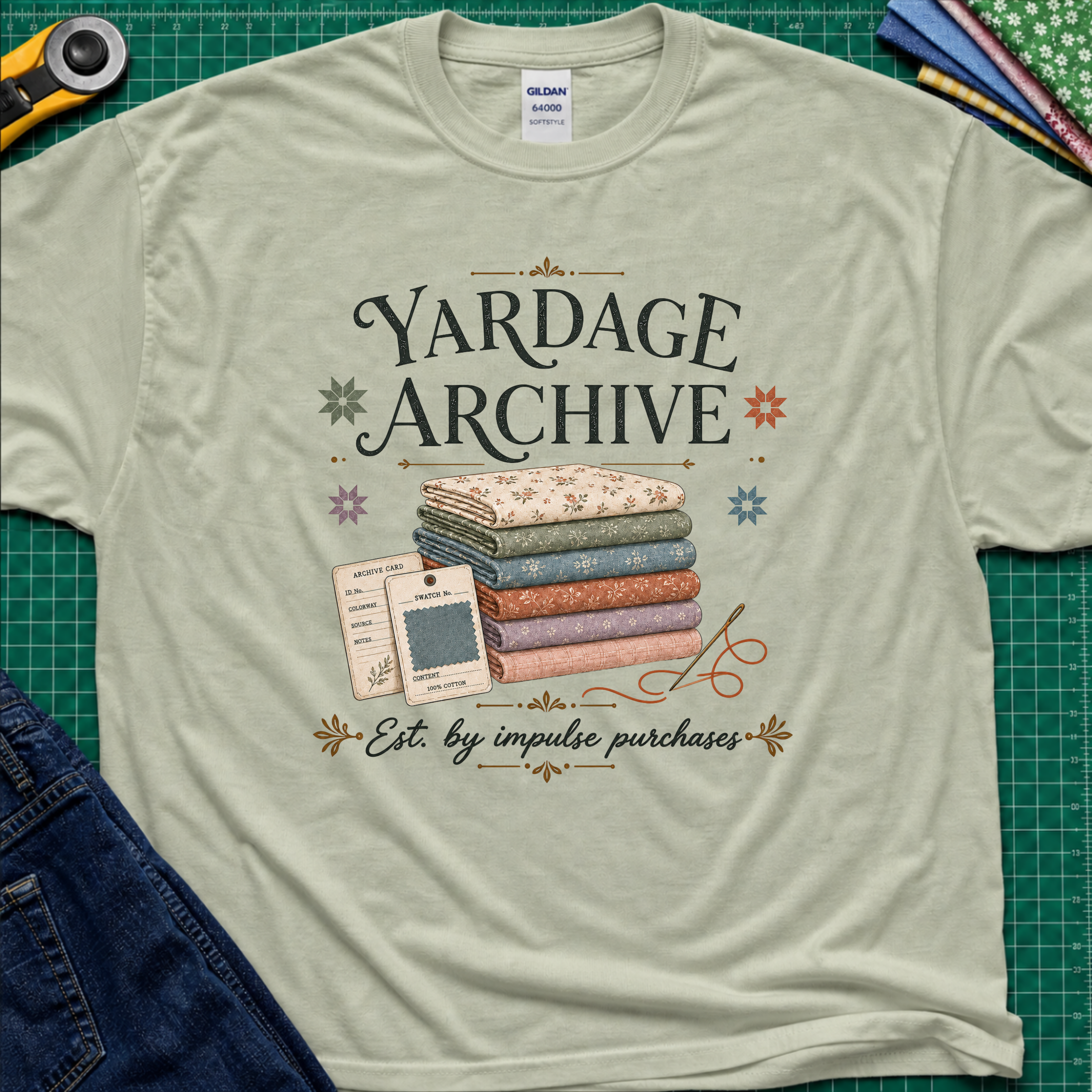A light sage green Gildan t-shirt laid flat on a green cutting mat, featuring a vintage-style graphic that reads 'Yardage Archive' with a stack of folded fabrics, a fabric label and swatch. Decorative craft tools and blue jeans are partially visible around the edges.
