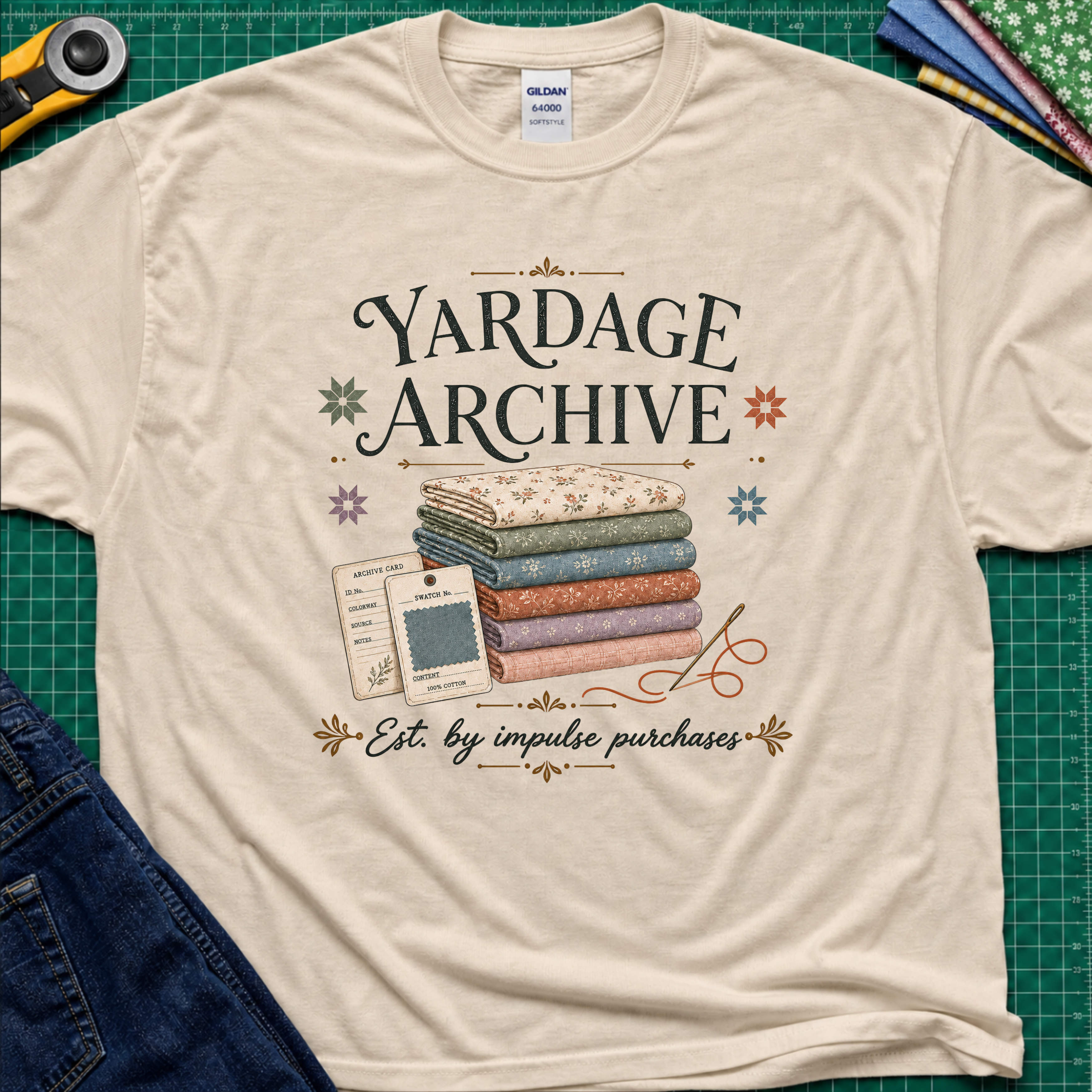 A dark cream Gildan t-shirt laid flat on a green cutting mat, featuring a vintage-style graphic that reads 'Yardage Archive' with a stack of folded fabrics, a fabric label and swatch. Decorative craft tools and blue jeans are partially visible around the edges.