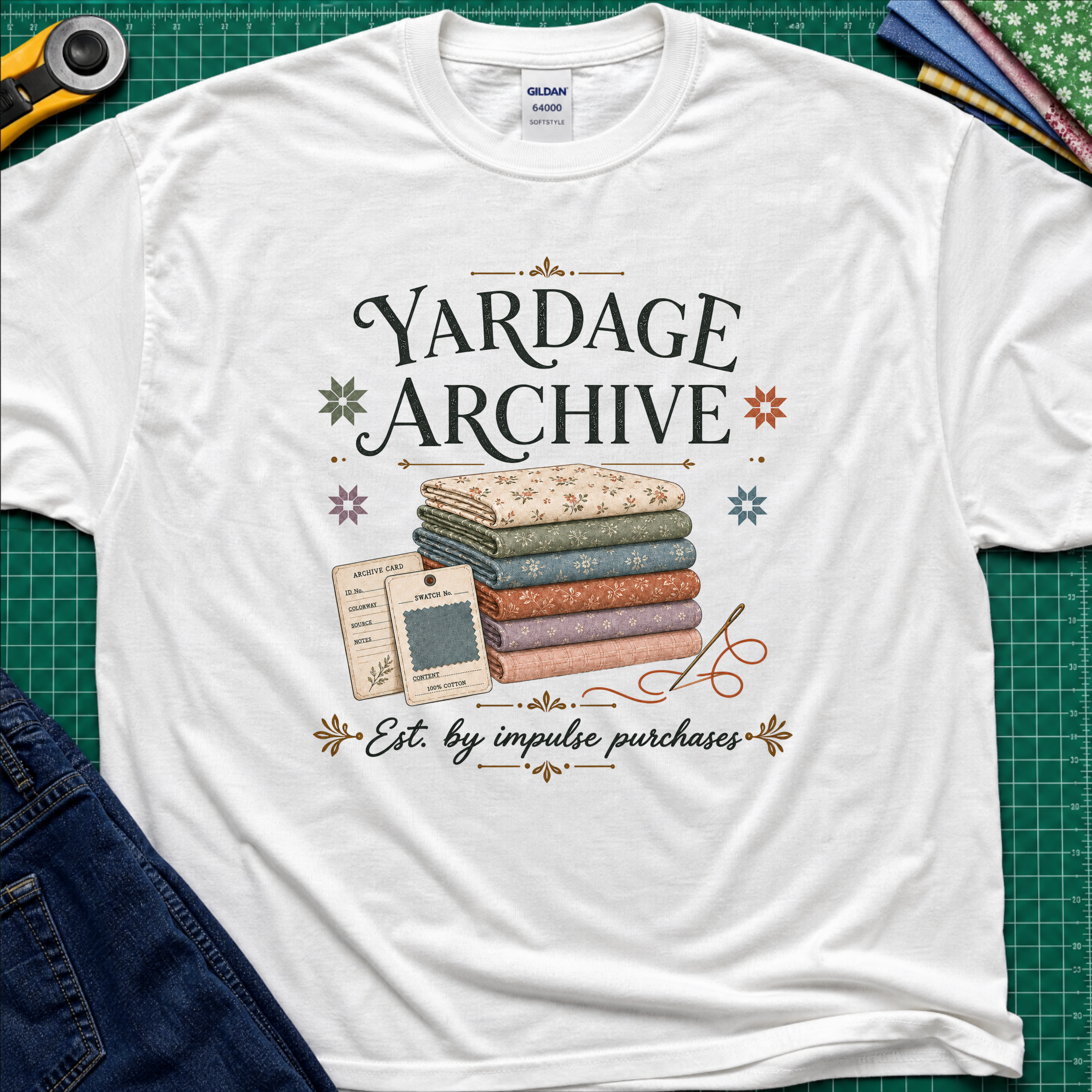 A white Gildan t-shirt laid flat on a green cutting mat, featuring a vintage-style graphic that reads 'Yardage Archive' with a stack of folded fabrics, a fabric label and swatch. Decorative craft tools and blue jeans are partially visible around the edges.
